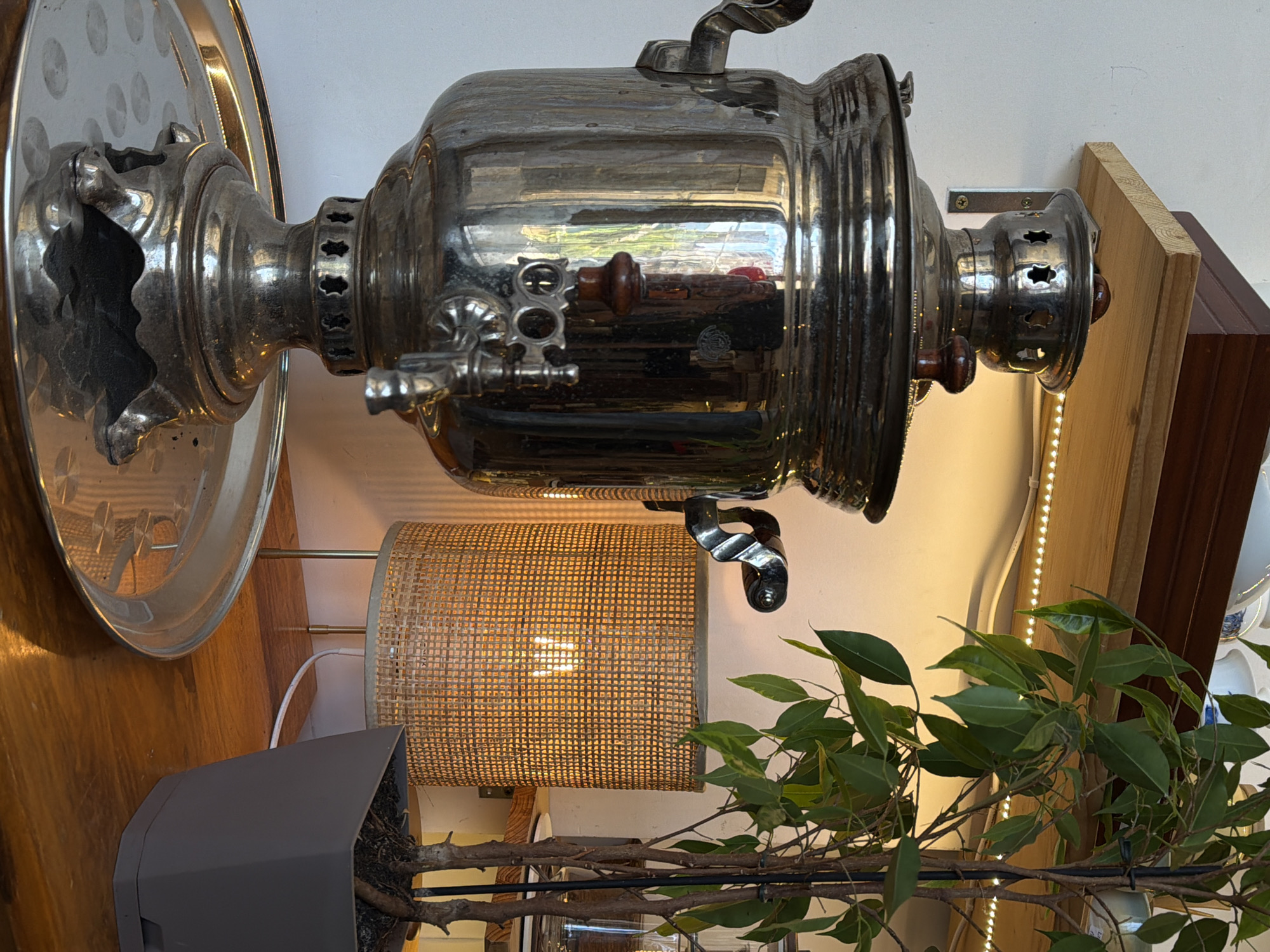 A close-up interior shot depicts a silver samovar, a traditional Russian metal container used for heating water, mounted on a light-colored wall. The samovar is accompanied by a circular silver tray. Below, there is a woven lampshade emitting a warm, yellow light. A potted plant with green foliage is visible, extending towards the right side of the frame. The background includes wooden panels and LED strip lighting. The location is Budva, Montenegro.