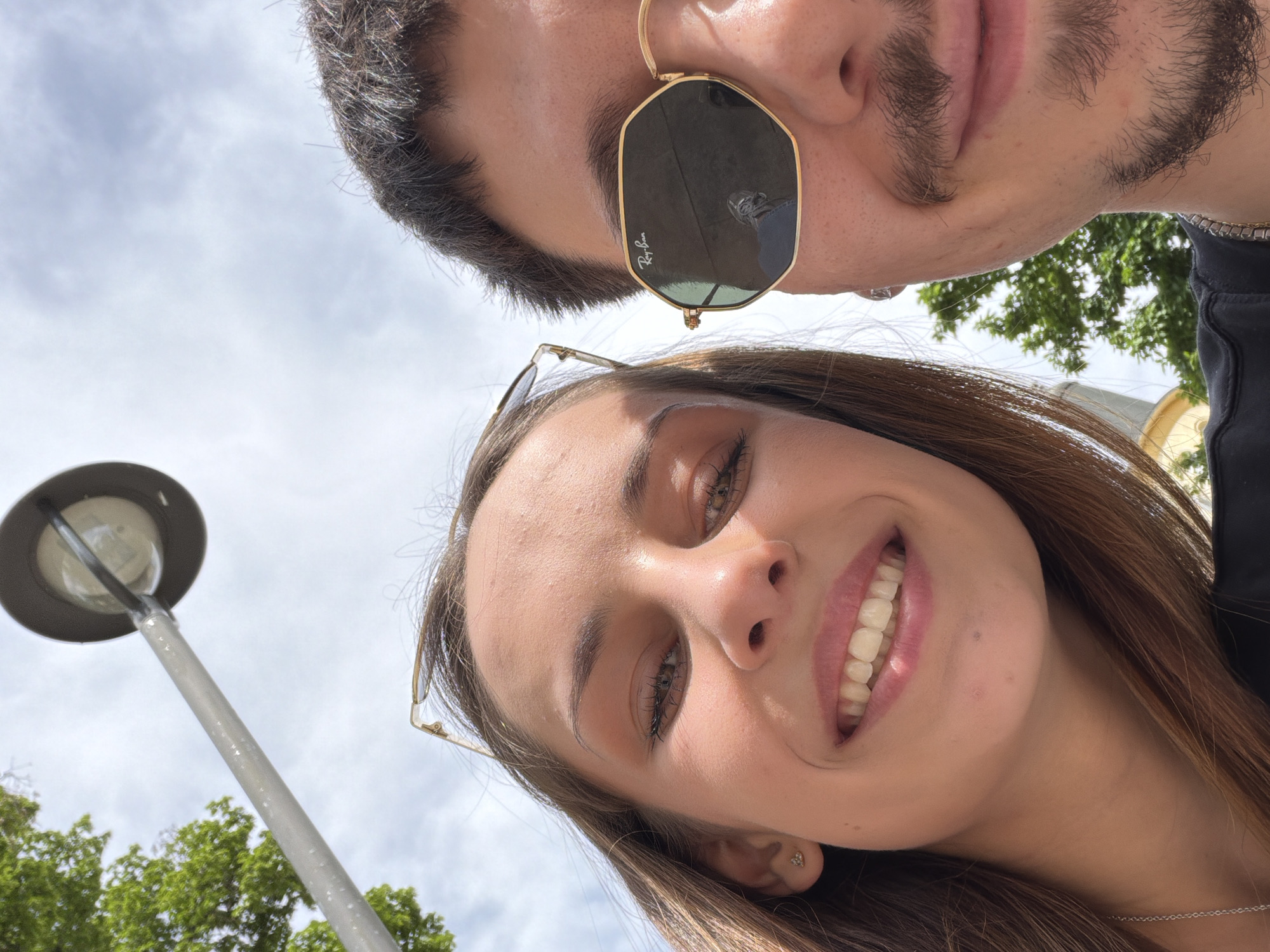A low-angle, close-up shot shows two people, a male and a female, likely taking a selfie. The male is positioned at the top, wearing gold-rimmed Ray-Ban sunglasses and sporting a beard. The female is positioned below him, smiling and also wearing glasses. Behind them, the background reveals a cloudy sky, trees, and a street light. The location is Sofia, Bulgaria.