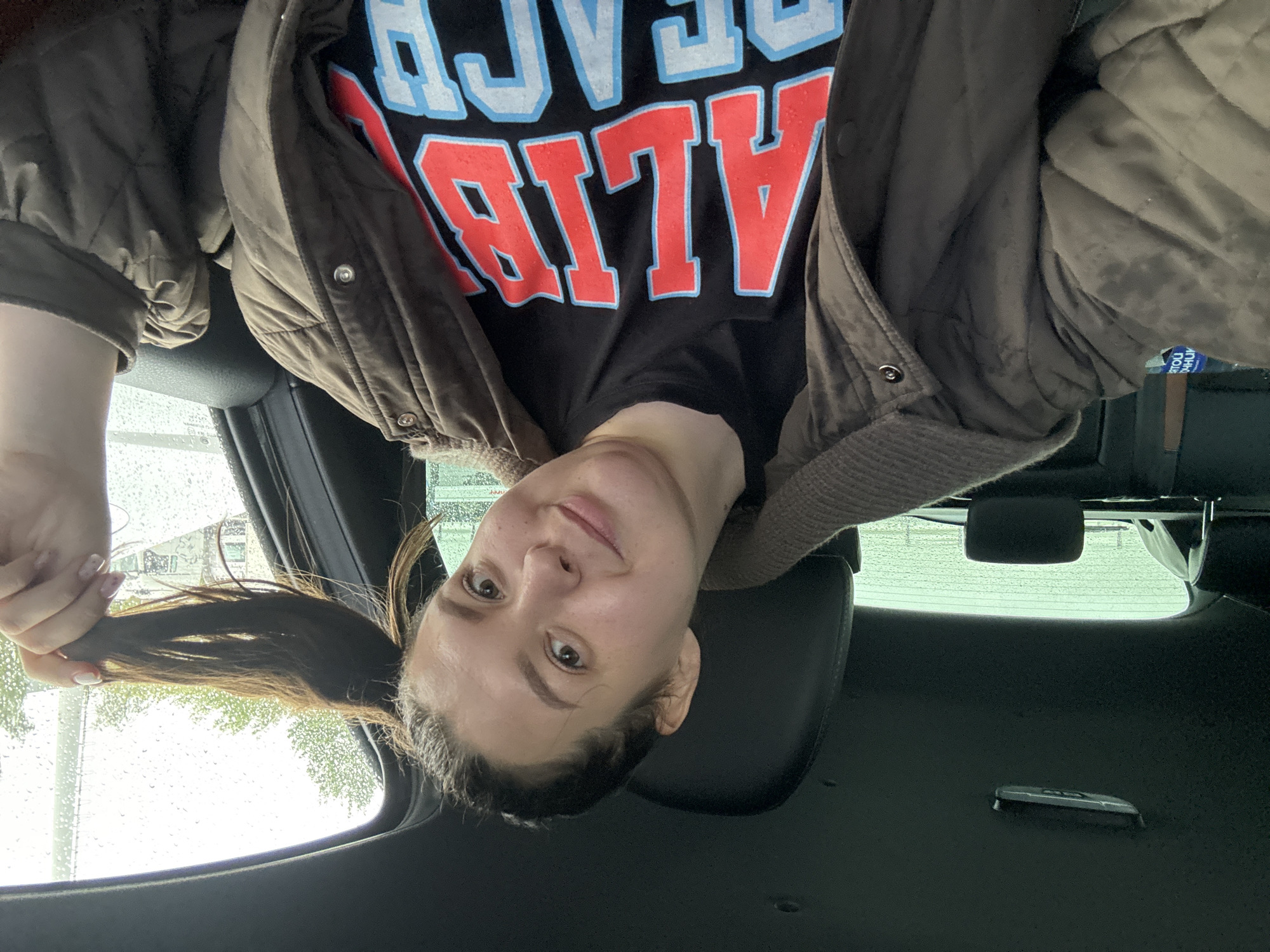 A woman is positioned upside down in the frame, likely inside a car. She wears a black t-shirt with "BEACH ALIVE" printed on it in red and blue letters, covered by a brown jacket. Her hair is pulled back into a ponytail, which she is holding in her hand. Raindrops are visible on the car window. The background shows a blurry view of the outside, making it difficult to determine the exact location. There is no information available that confirms the location in the city of Izhevsk, Russia.