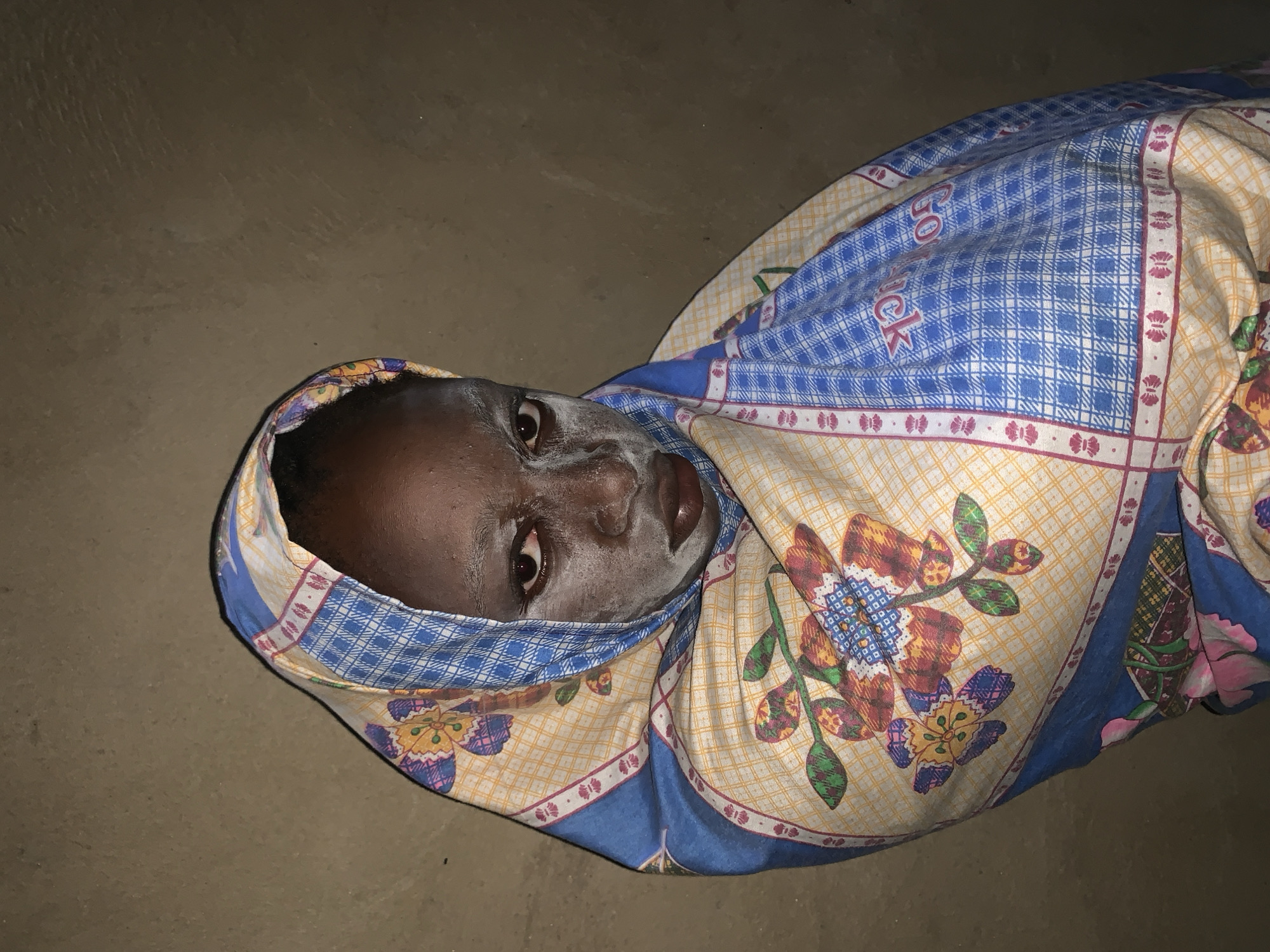 A person with dark skin and what appears to be white powder on their face is wrapped in a patterned cloth. The cloth is predominantly blue and white plaid with a border featuring floral designs in various colors including yellow, orange, and blue. The person is lying on a surface that appears to be a floor made of a concrete-like material in a dimly lit area. The photo appears to be taken in Agban, Nigeria. The person's eyes are open, and their expression is neutral. The blanket is drawn up around their head.