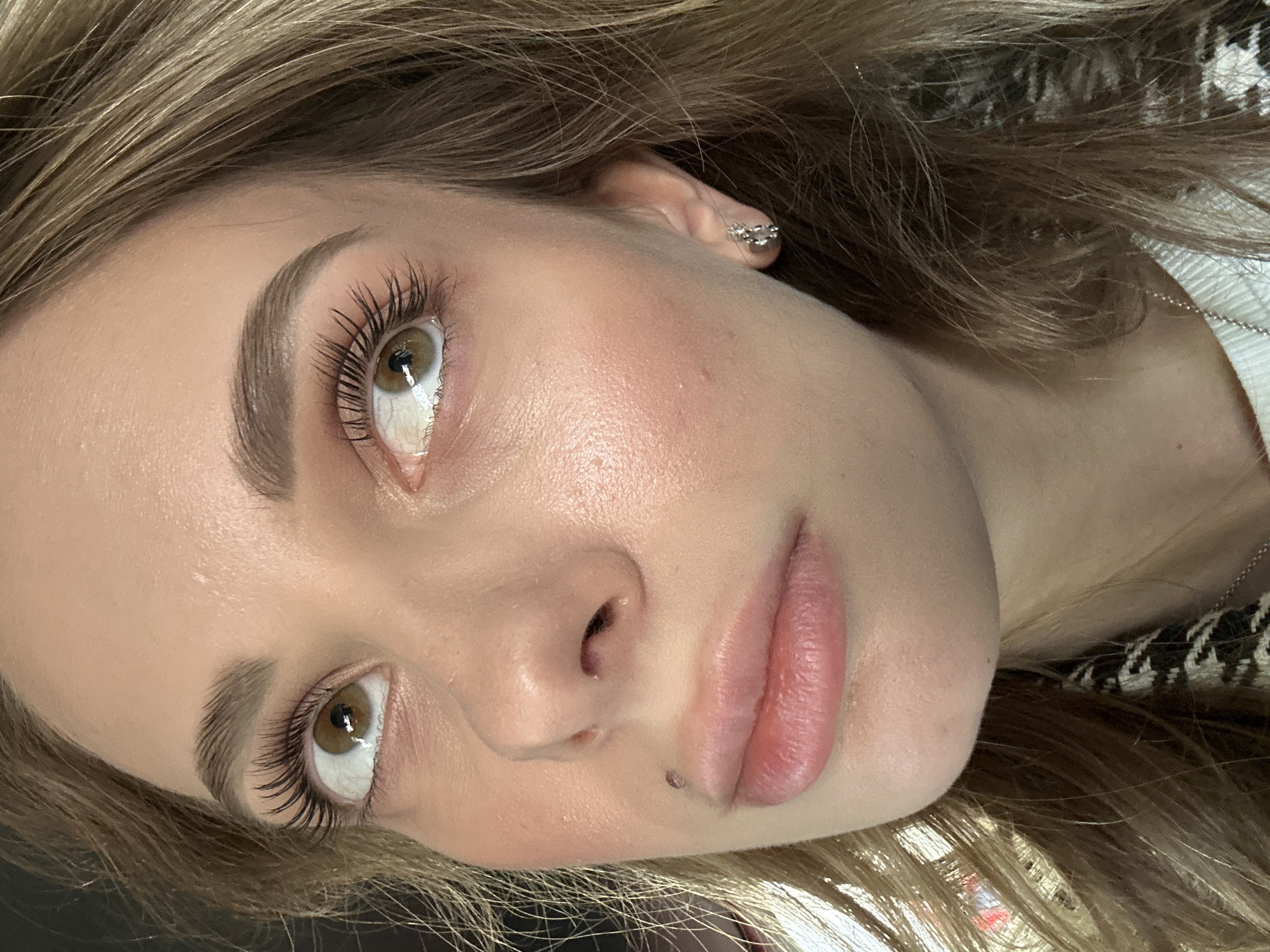 A close-up, upward-angled shot shows a woman's face with light brown hair. Her eyes are hazel, and she has long eyelashes. She has full lips and fair skin. A silver earring is visible in her left ear. A mole is present near her lower lip. A silver necklace is visible near her neck. The woman appears to be wearing makeup. The image was likely taken indoors, given the lighting and context. There is no evidence in the image to suggest it was taken in Warsaw, Poland.