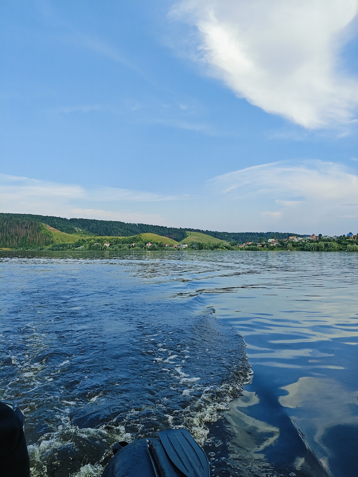 This is a medium shot, outdoor, horizontal photograph taken from a boat on a body of water, likely a river, in the city of Noviy, Russia. The water in the foreground is dark blue and choppy, with white foam and bubbles trailing behind the boat, indicating movement. The right side of the frame shows the undisturbed, smooth surface of the water, reflecting the sky and clouds.
In the background, a lush green landscape with rolling hills extends across the horizon. Dense forests cover the upper slopes of the hills, with lighter green patches of grass and scattered trees lower down. A small village is nestled along the shoreline, with several houses visible amongst the trees, some with red roofs. The sky is a clear, bright blue with a few wispy white clouds.
The lower left corner of the image shows a portion of the boat, including what appears to be an oar or paddle. No people are visible in the frame. The overall atmosphere is serene and natural, suggesting a peaceful day on the water.