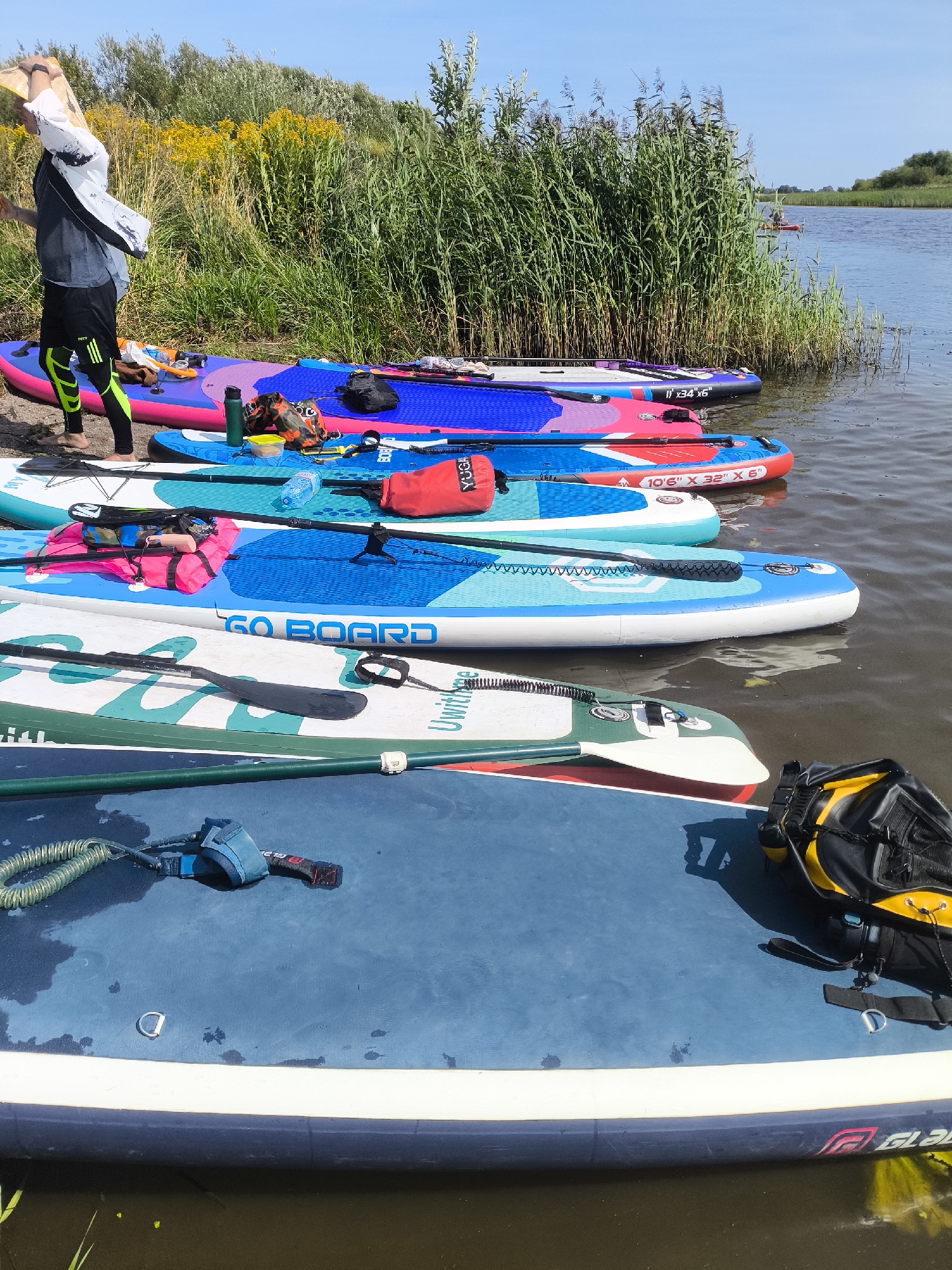 The image shows a collection of stand-up paddleboards (SUPs) lined up on the shore of a body of water, likely a river or lake, with tall reeds growing along the bank. In the background, a person is visible in a red kayak on the water. The foreground features several SUPs of various colors and designs, some with paddles and leashes attached. A person is standing on the shore, partially visible, wearing athletic clothing and holding what appears to be a yellow object over their head. The environment is sunny with a clear blue sky. The scene suggests preparation for or the aftermath of a paddleboarding activity. The location is identified as Ushakovo, Russia, though this information is not visually discernible from the image itself.
