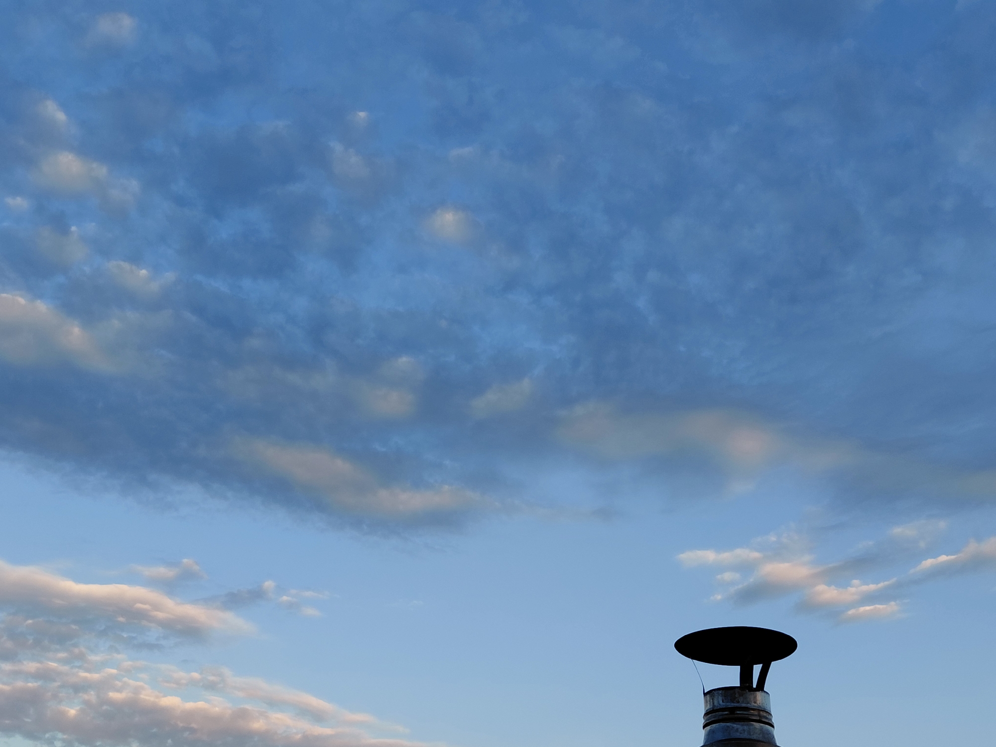This image, captured in Verhnyaya Podstyopnovka, Russia, features a wide-angle view of a partially cloudy sky during daylight hours. The sky is a vibrant blue, with a scattering of wispy white and gray clouds. In the lower right corner of the frame, a portion of a chimney is visible, topped with a black cap. The chimney appears to be made of metal, with visible rings. No people or animals are present in the scene. The overall impression is one of a calm, overcast sky over a rural or suburban landscape.