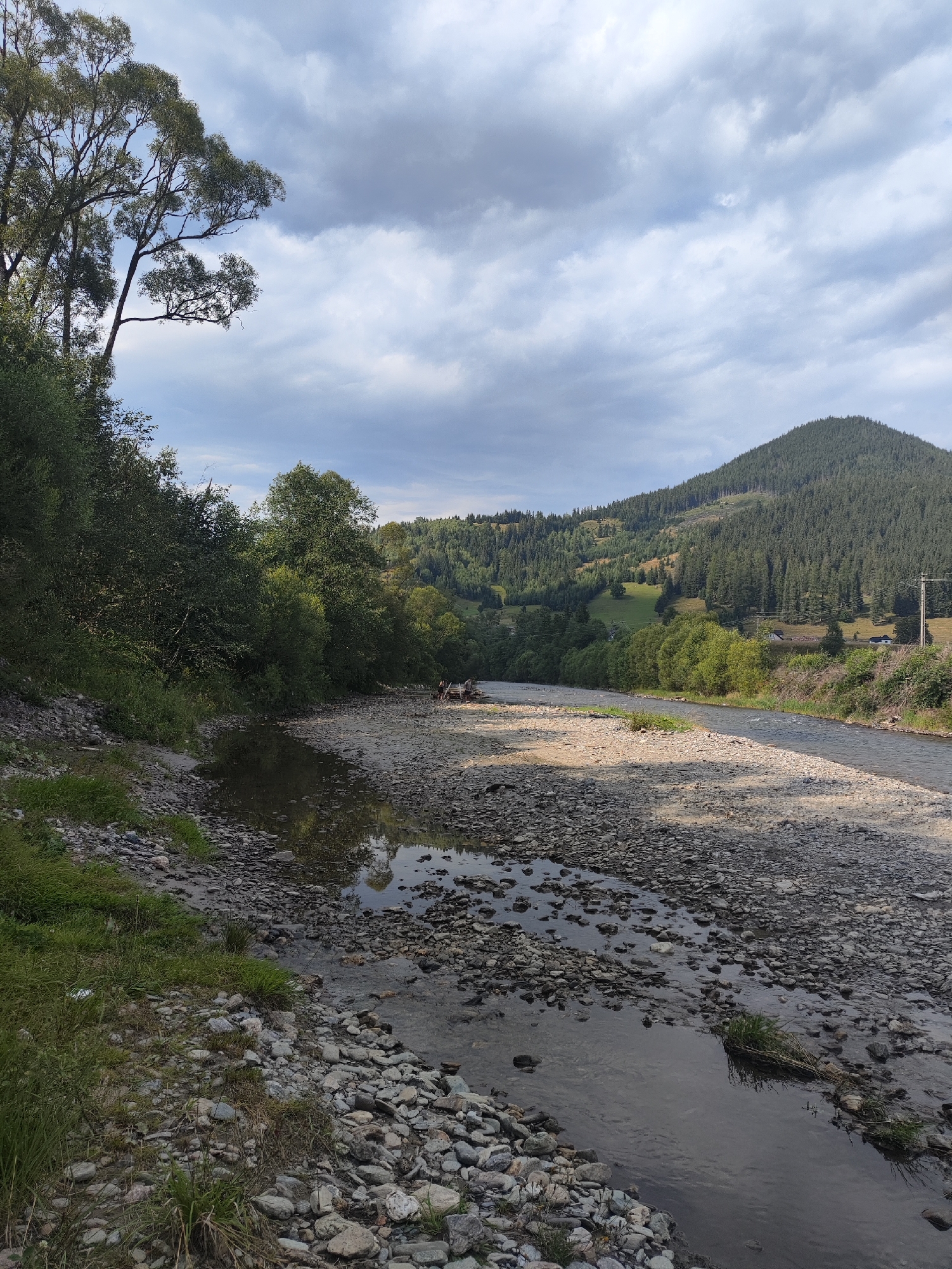 The image depicts a wide, rocky riverbed with a shallow stream flowing through it. The riverbanks are lined with lush green trees and vegetation. In the background, a large, forested mountain rises under a cloudy sky. A few small structures and utility poles are visible on the mountainside. In the foreground, there are a few people sitting by the river. The scene appears to be a natural landscape, possibly a park or a rural area, but no specific location in Botoș, Romania, can be identified from the image.