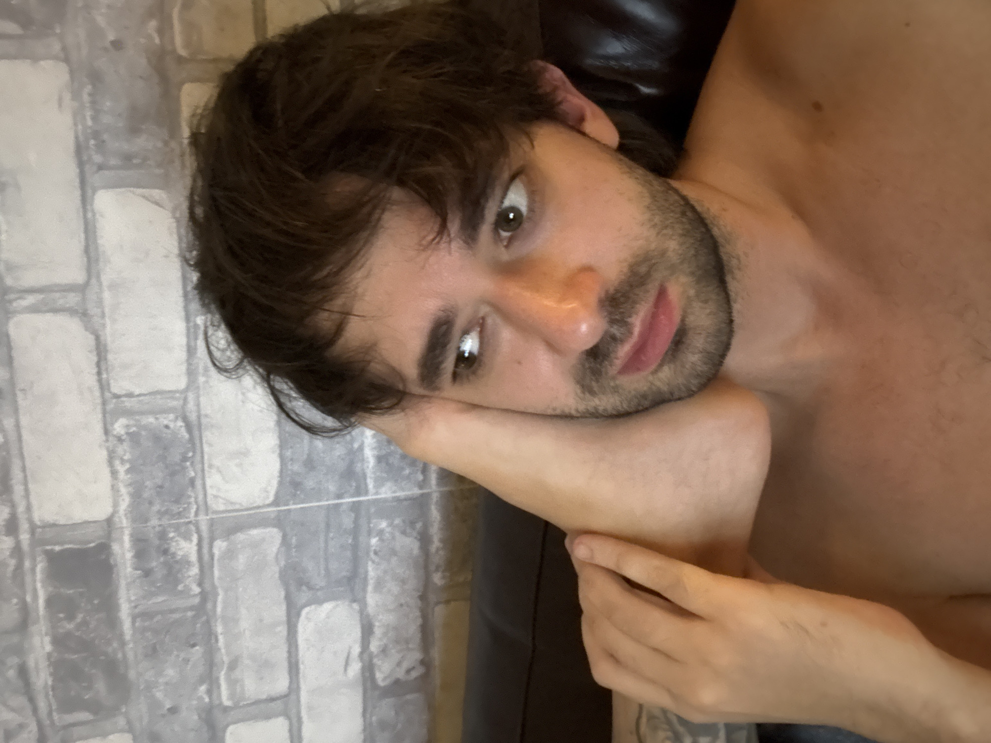 The image shows a close-up of a man's head and upper body. He has dark brown, messy hair and a beard. His eyes are looking to the left. His left arm is bent, and his foot is placed against the side of his face, obscuring part of his cheek and jawline. The background appears to be a brick wall. No specific location in Moscow, Russia, can be identified from the image.