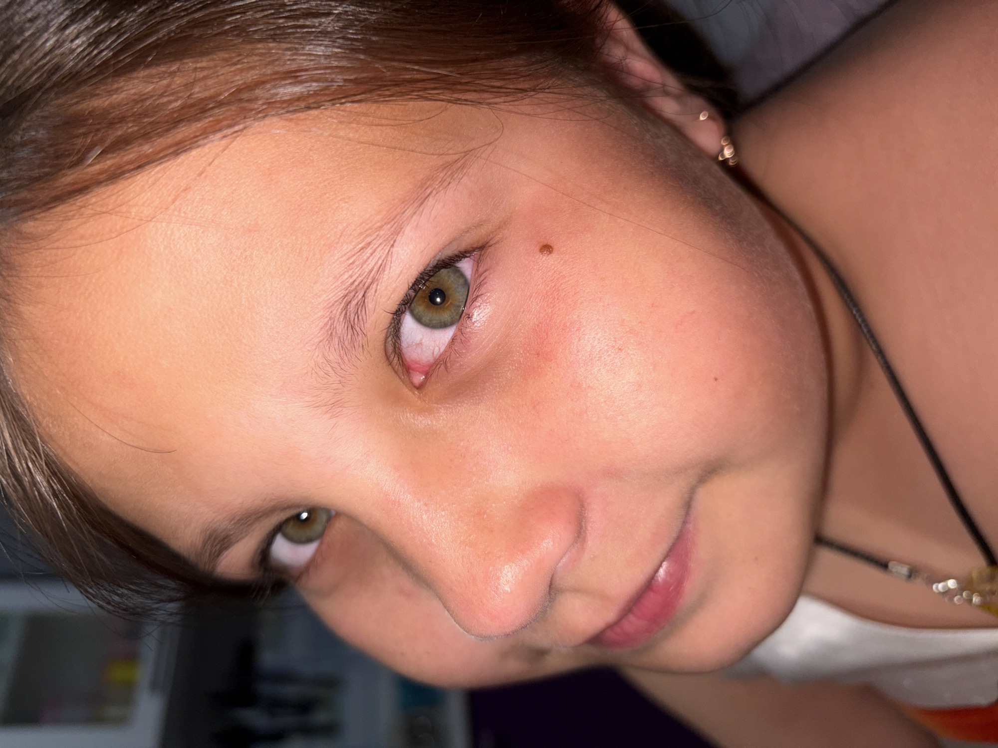 A close-up shot of a young girl with fair skin, brown hair, and green eyes looking up at the camera with a slight smile. There's a small mole visible near her right eyebrow. She is wearing a necklace with small beads. The image doesn't contain sufficient details to confirm a location in Novopokrovskaya, Russia.