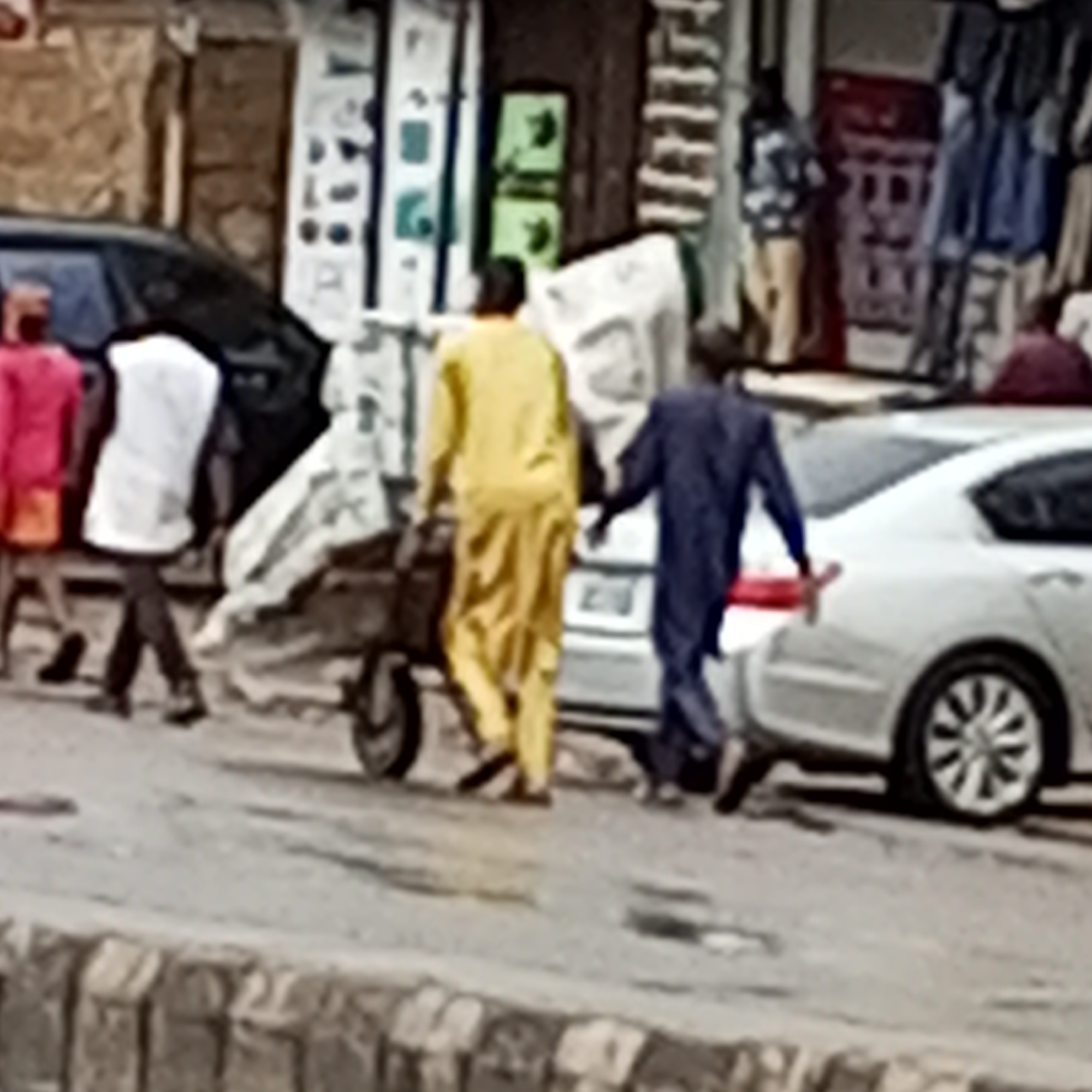 In Maiburiji, Nigeria, a street scene unfolds with several individuals and vehicles. A black SUV is parked on the left, with a person in a bright pink top and orange bottoms visible behind it. Next to them, a person in a white shirt and dark pants walks away from the viewer, pushing a cart. To the right, two men walk together. One is dressed in a bright yellow garment, and the other wears a dark blue traditional outfit. They are walking beside a white sedan, which has its rear passenger-side door open. The background features storefronts with various items displayed, including clothing hanging outside. The ground is paved, and a stone barrier runs along the bottom of the frame.
