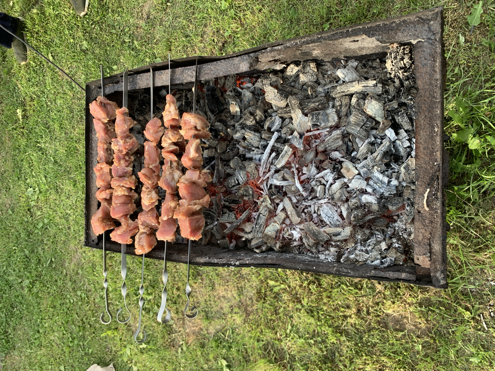 An outdoor, high-angle view captures a rustic, rectangular metal brazier positioned directly on green grass within Відзаўскі сельскі Савет, Belarus. Inside the brazier, a bed of glowing red charcoal mixed with white ash is visible. Four metal skewers, each designed with a distinctive twisted handle, are horizontally suspended across the top of the brazier. Each skewer holds multiple pieces of raw, cubed, marinated meat, consistent with pork or chicken, positioned directly above the hot coals. The metal structure of the brazier exhibits signs of wear, charring, and minor rust along its edges. In the upper right section of the image, the lower legs and feet of an individual are visible, wearing dark trousers and olive-green shoes. A slender, dark metal rod extends from the approximate position of this person towards the brazier. A small, light-colored object rests on the grass in the upper left quadrant. The scene is illuminated by natural daytime light.