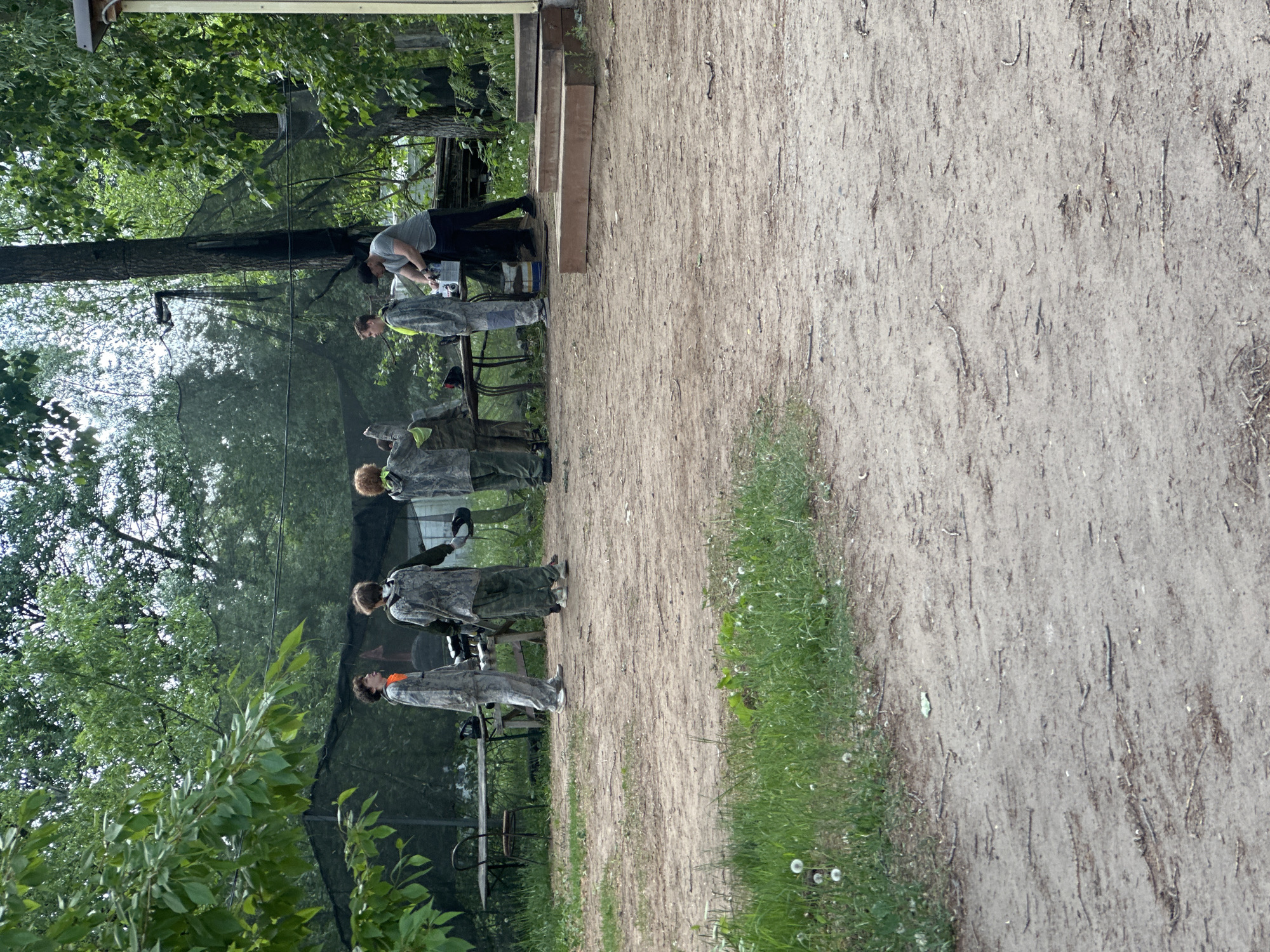 The image shows a group of people lined up outside at a paintball field. The location appears to be a wooded area with a dirt path. Five people are standing in line wearing camouflage paintball outfits and protective masks, facing a table where an individual is filling paintball hoppers. To the left of the line of people is foliage, trees, and a net. The right side shows the dirt path, some grass, and dandelions. The scene takes place outdoors. There is no specific evidence indicating Chaikovsky, Russia.