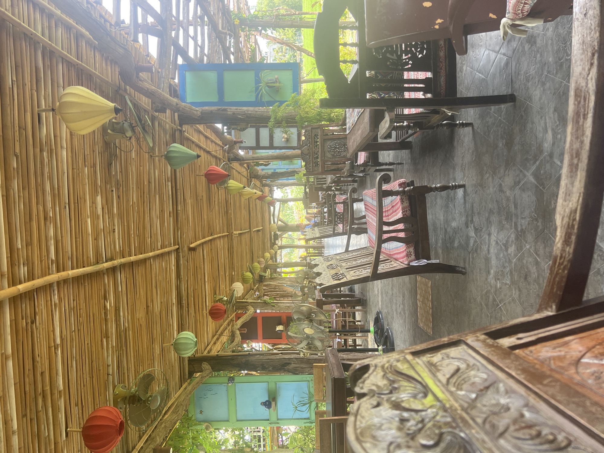 This image depicts the interior of an open-air restaurant in Hoi An, Vietnam, likely designed in a traditional style. The ceiling is constructed from natural bamboo, and several colorful lanterns (red, yellow, and green) are suspended from it, along with vintage-style electric fans. The seating area features numerous wooden tables and chairs, many of which appear to be antique or handcrafted, with carved details and fabric cushions.
In the background, tall green fields are visible through the open structure, suggesting the restaurant is situated in a more rural or scenic area of Hoi An, possibly near rice paddies. Two large, ornate wooden doors painted in vibrant blue and green hues are mounted on posts, serving as decorative elements and framing views into the surrounding greenery. Several electric fans are positioned throughout the seating area.
There are no discernible people actively engaged in dining or service in the foreground or midground. The overall impression is one of a tranquil, rustic dining space designed to offer a relaxed ambiance, possibly with an emphasis on local aesthetics. The lighting suggests it is daytime, with natural light filtering through the bamboo roof and open sides.