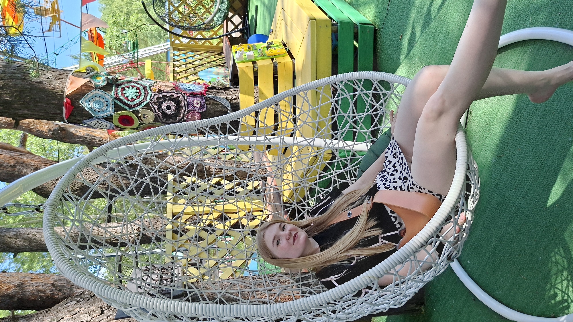 A young blonde woman with long hair is lying in a white wicker hanging chair outdoors. She is wearing a black top and a white and black leopard print bikini bottom. Her legs are crossed and extended upwards. The hanging chair is suspended from a tree branch, and the background consists of a tree trunk, green grass, and a colorful structure made of yellow and green wood. There are also crochet decorations hanging on the tree and colorful flags in the upper left corner. The scene appears to be set in a park or a backyard during the daytime, with bright sunlight casting shadows. The woman has a relaxed expression. No visible text is present in the image. The location is identified as Chaikovsky, Russia.