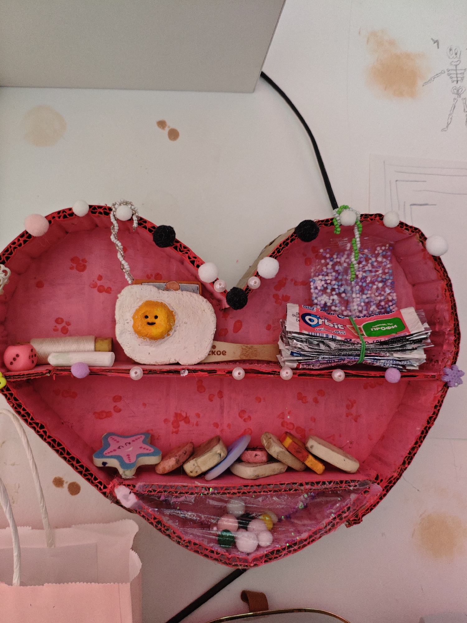 The image shows a handmade heart-shaped shelf mounted on a white wall. The shelf is decorated with pink paper and red trim. Inside the shelf are various small objects. On the top shelf, there is a decorative egg with a smiley face, a lipstick, a pink die, and a stack of gum wrappers tied with a green rubber band. Above the gum wrappers, there are small colorful beads in a clear plastic bag. On the bottom shelf, there is a blue and pink starfish-shaped object, several small oval-shaped objects, and a clear plastic pouch filled with colorful beads and small white pom-poms. The wall also has some brown smudges and a sketch of a skeleton. The overall scene appears to be an interior setting. There is no identifiable information to locate this scene in Трудовское сельское поселение, Ukraine.
