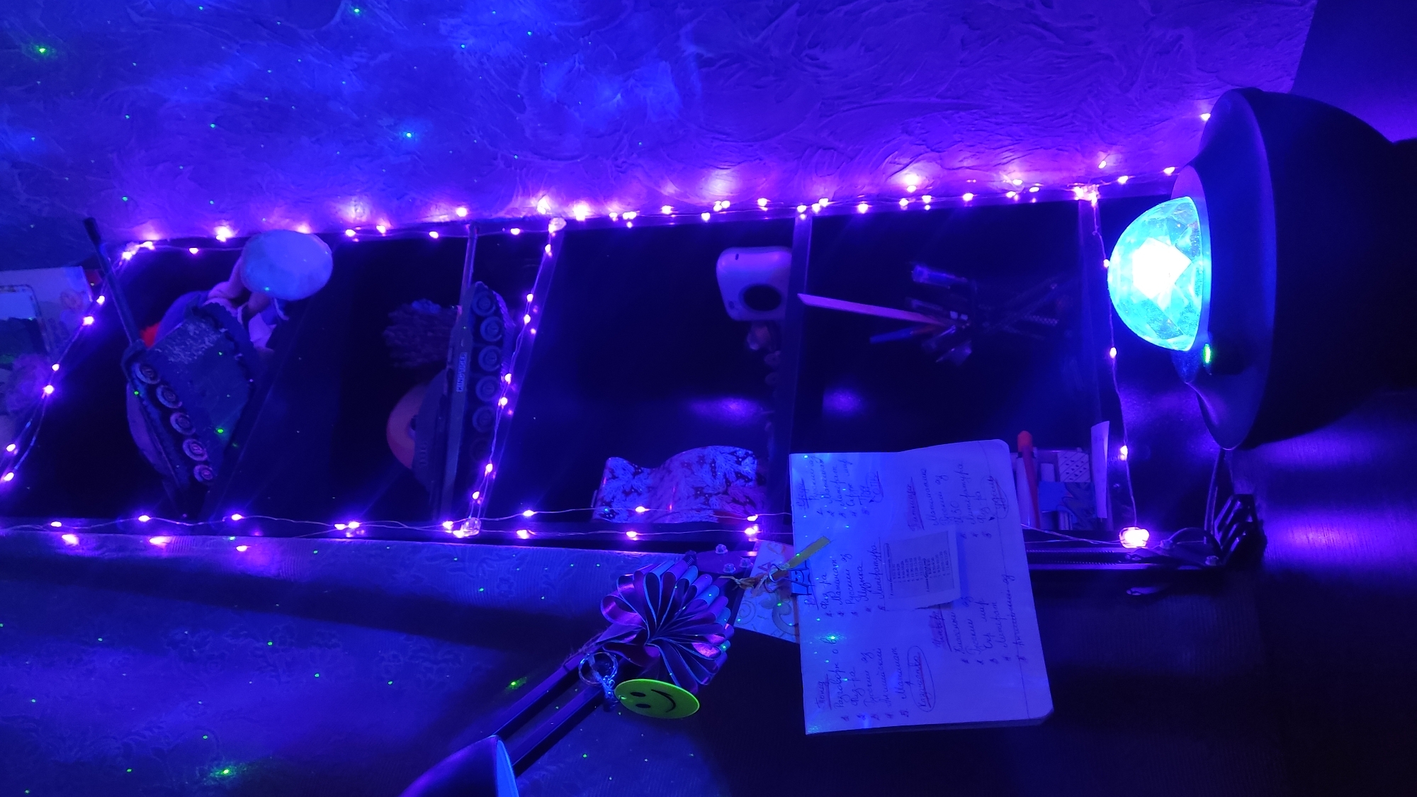 The image is taken indoors, illuminated by blue and purple LED lights. The scene appears to be a shelf or display area, possibly a child's room. On the shelves, there are toy tanks, a white globe or balloon, and other miscellaneous objects. In the foreground, a notebook with handwritten notes in Russian is visible, along with a pen, a decorative ribbon, and a smiley-face keychain. A bright blue light source is positioned on the right side of the image. The OCR data from the notebook suggests the location is in Russia and contains references to school subjects. Given the reference to "Russian language", it is reasonable to infer that the location is in the city of Krasnodar, Russia, due to the language.