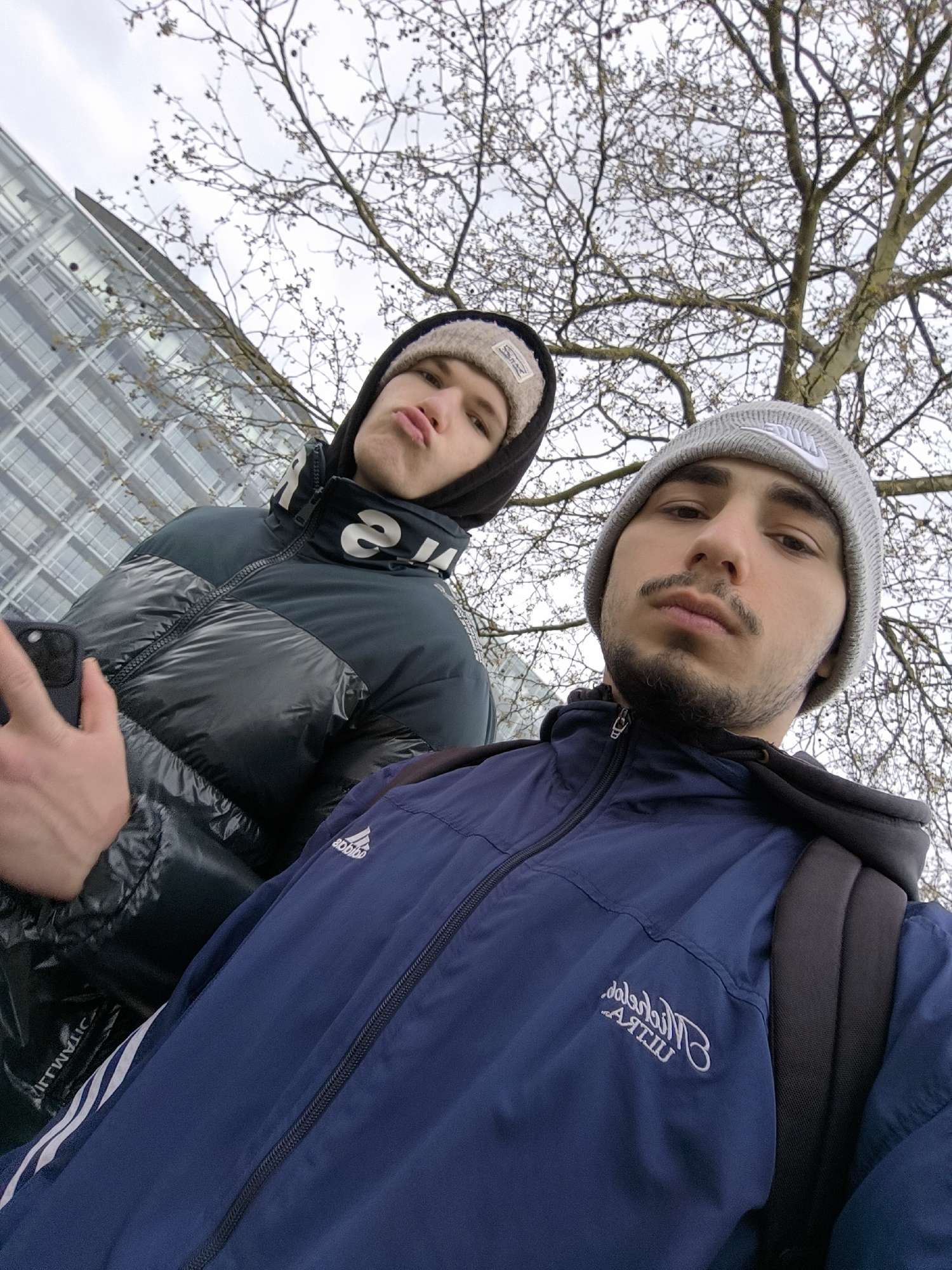 Two young men are posing for a selfie outdoors. The man in the foreground wears a navy blue athletic jacket with a hood pulled down, a gray knit beanie with a visible Nike logo, and a dark backpack. He has a short beard and mustache. The man behind him wears a shiny, dark puffy jacket with a hood pulled up, revealing a light-colored knit hat underneath. He is making a pouting face. A modern building with many windows is visible in the background, along with a tree with bare branches. The weather appears overcast. Based on visual markers, the location is Strasbourg, France.