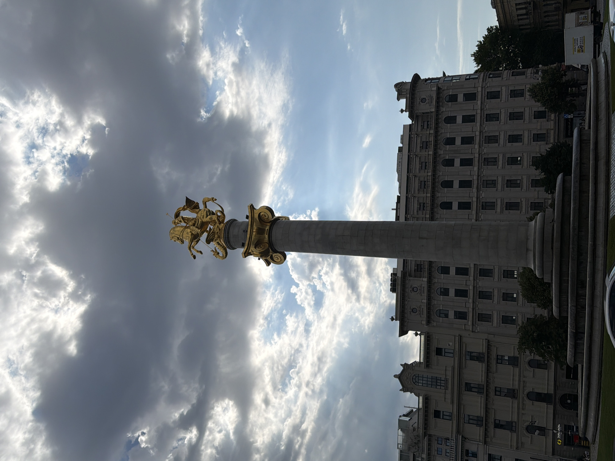 A golden statue of Saint George on horseback, spearing a dragon, sits atop a tall, gray stone column in Freedom Square, Tbilisi, Georgia. The statue is illuminated by the sun breaking through dramatic, dark clouds. Behind the monument, a large, ornate building with numerous windows stands out against the sky. The lower part of the image shows the circular base of the monument and a hint of a fountain. Trees and other buildings are visible in the background. The overall scene suggests a dramatic and possibly overcast day in a prominent urban location.