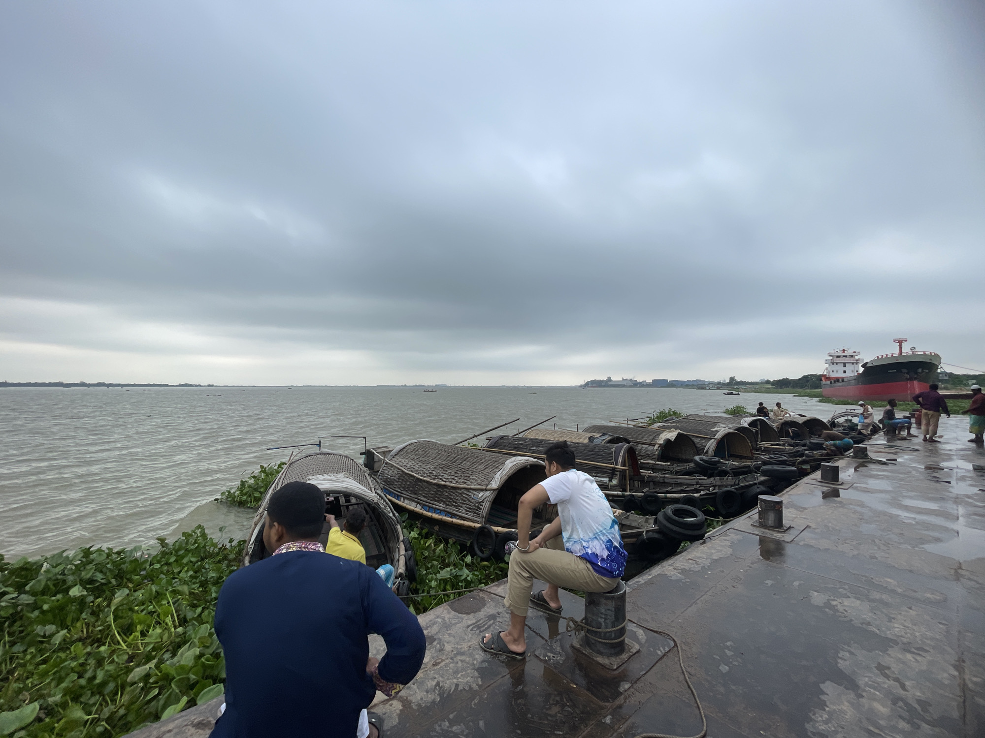The scene shows a waterfront in Sonargaon, Bangladesh, under a cloudy sky. The water is choppy and dotted with small boats. On a concrete pier, multiple small, covered boats, presumably for passenger transport, are docked and entangled with water hyacinth.
Several people are visible: Two men are prominent in the foreground. One, wearing a blue shirt and a black cap, stands with his back to the viewer. Another, wearing a white shirt and khaki pants, sits on a mooring bollard. Further along the pier, more people are present, engaging in activities like sitting and standing. A large ship with a red and white hull is docked at the pier as well. The pier surface appears wet, likely due to recent rain or proximity to the water.