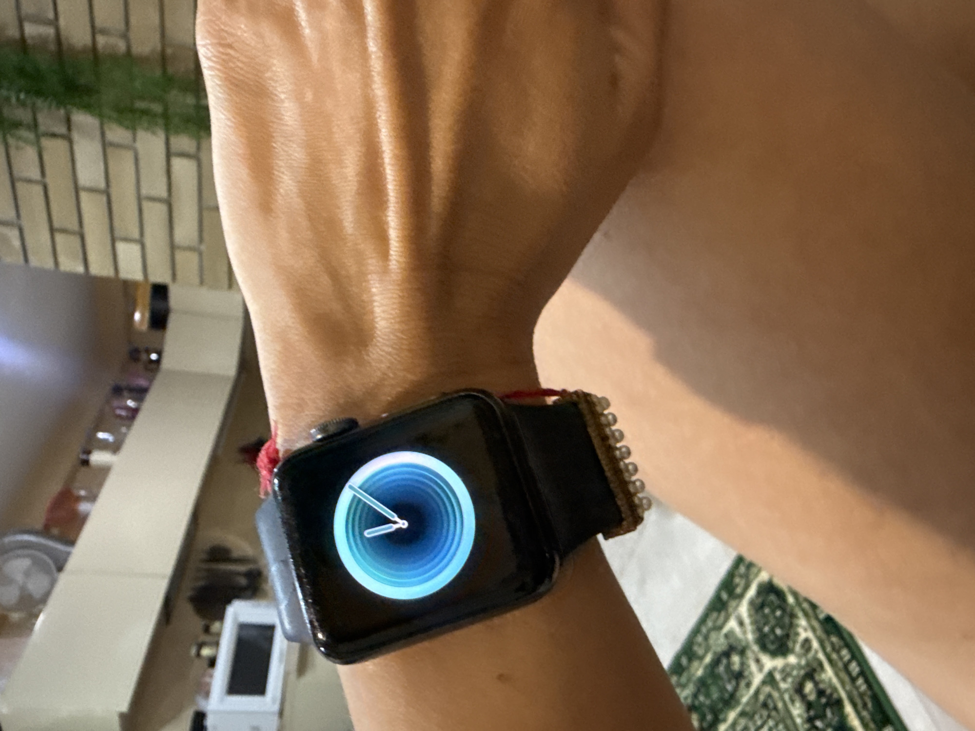 A close-up shot focuses on a person's left wrist wearing a black Apple Watch with a blue and white analog watch face. The watchband is black, and a red string is visible tied to it, along with decorative beads. The background shows a brick wall and cabinetry, suggesting an indoor domestic setting. The lighting is warm and artificial.