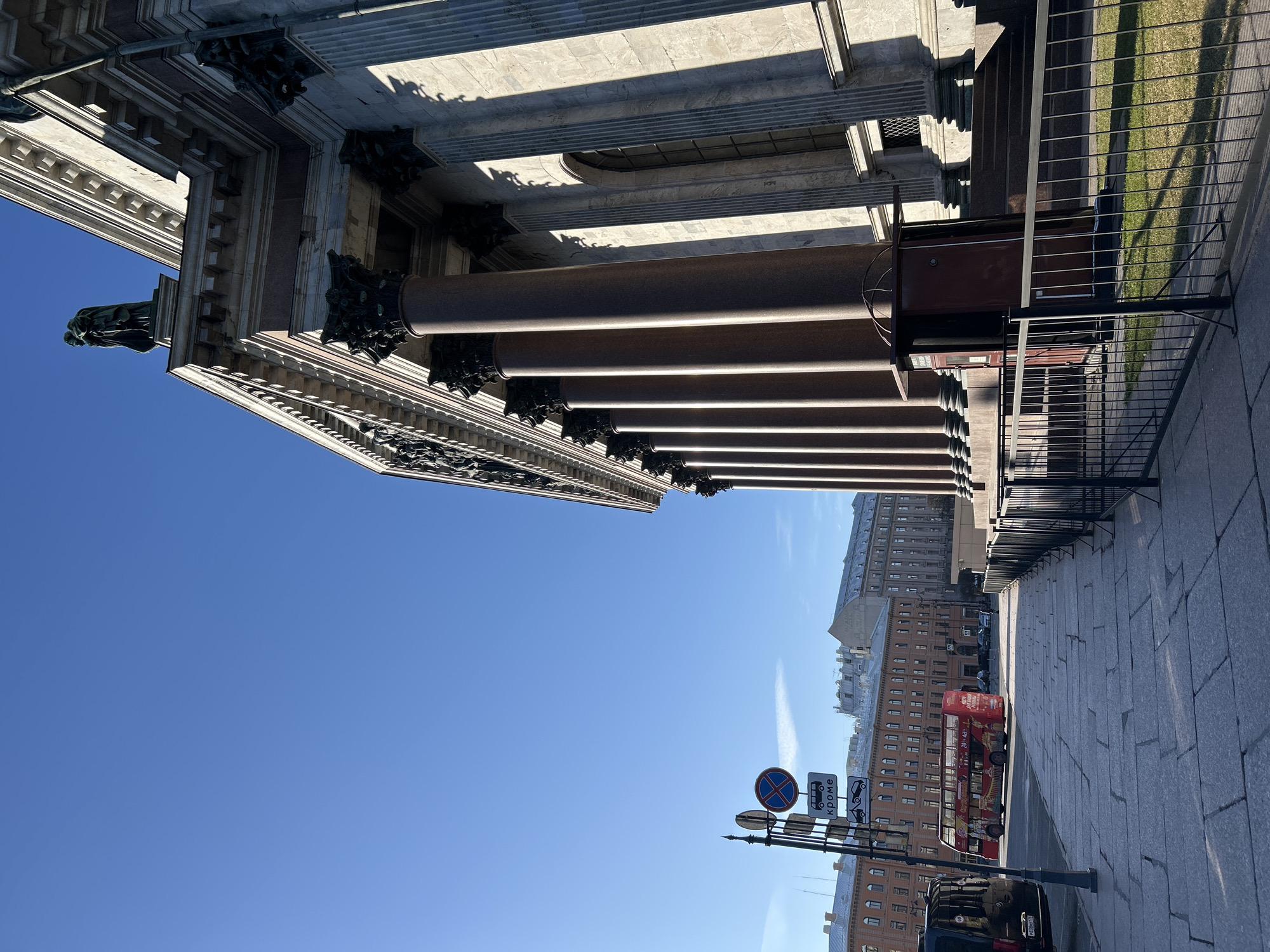 The image depicts a view of Saint Petersburg, Russia, featuring the colonnade of St. Isaac's Cathedral on the left side of the frame. The sky is clear and blue. On the right side, there are buildings, a red double-decker tour bus, traffic signs, a street lamp and part of a parked black car. The pavement is made of granite stones.