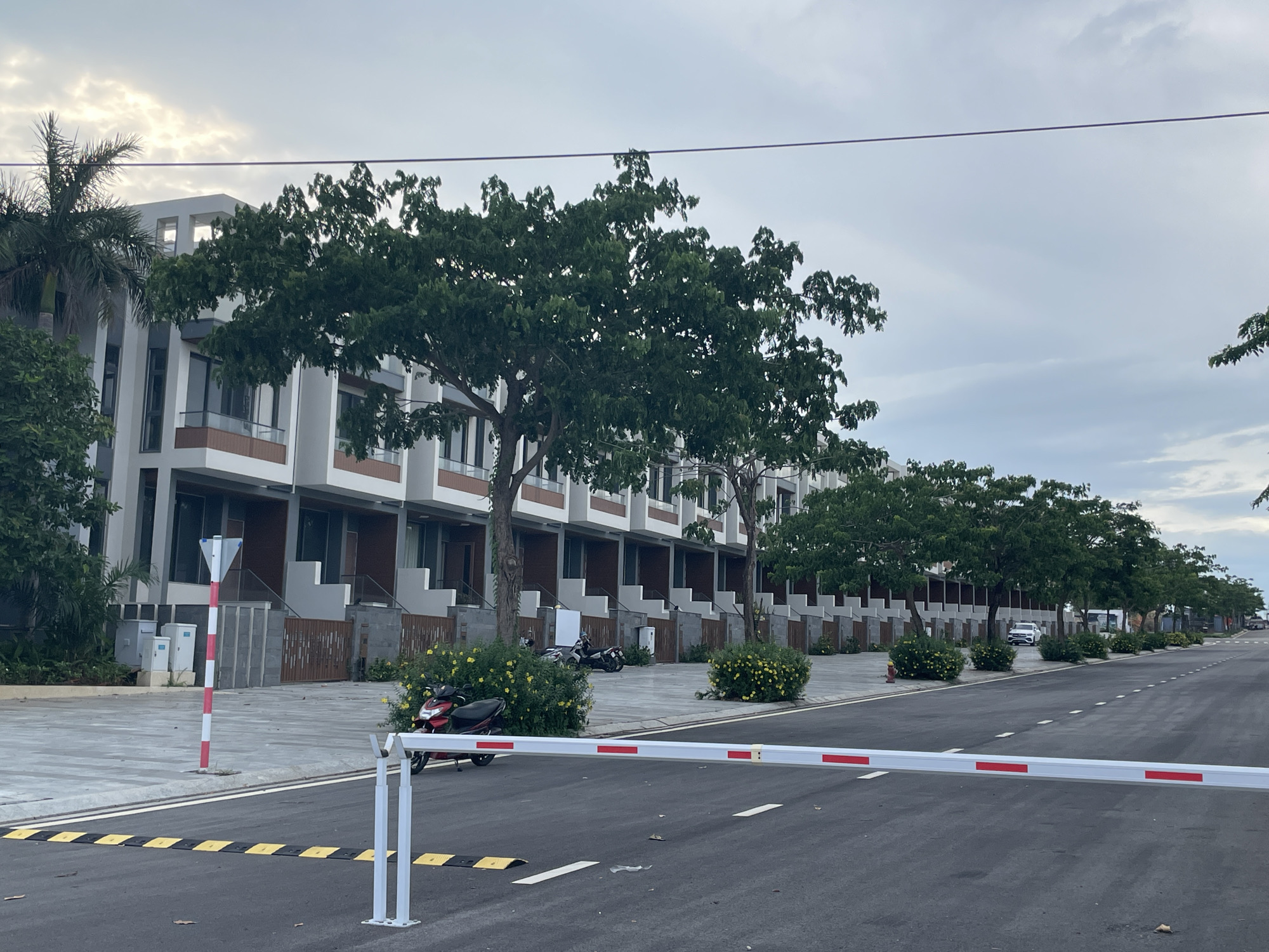 The image displays a modern residential street in Phu Thuy Ward, Vietnam, under an overcast sky. The scene is dominated by a long row of contemporary multi-story townhouses extending into the distance on the left side of the road. These residences feature a consistent modern architectural style with white and grey facades, large windows, and some brown-toned accents on balconies and exteriors. Each house appears to have a small front yard.
In the foreground, a white and red striped automated barrier arm stretches across an asphalt road, which is marked with white lines and black and yellow speed bumps. To the left of the barrier, a red and black scooter is parked on the sidewalk, nestled among yellow flowering bushes. Further down the street, additional motorcycles are visible parked among similar vegetation, and a white car is parked on the right side of the road.
The street is lined with numerous large trees with dense green foliage, providing shade and partially obscuring the lower levels of the townhouses. A tall palm tree is visible on the far left. Other street furniture includes a red and white striped pole with a triangular white sign on top, two white utility boxes near the barrier on the left, and a red fire hydrant further down the street. A single thin, dark overhead wire spans horizontally across the upper portion of the cloudy sky. No people are present in the scene. The overall impression is of a well-maintained, newly developed residential area.