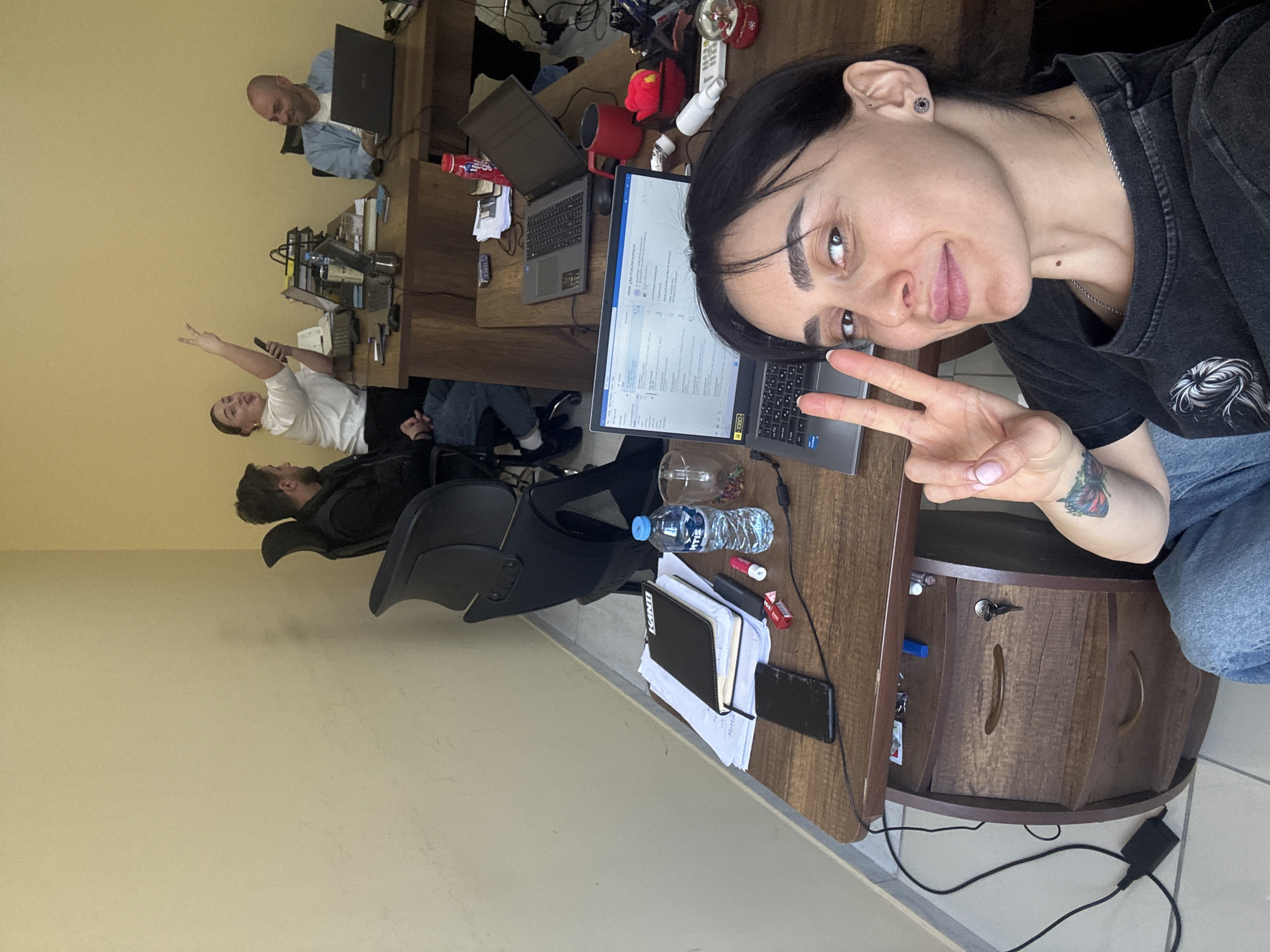 The image shows a woman taking a selfie in an office environment. She is making a peace sign with her hand. Behind her, other people are working at desks, including a bald man working on a laptop. There is a laptop, a glass of liquid, a water bottle, and a notebook on the desk in front of the woman. The room has wooden desks and a beige wall. The location is likely Tbilisi, Georgia, based on the context of the image and the presence of other individuals.