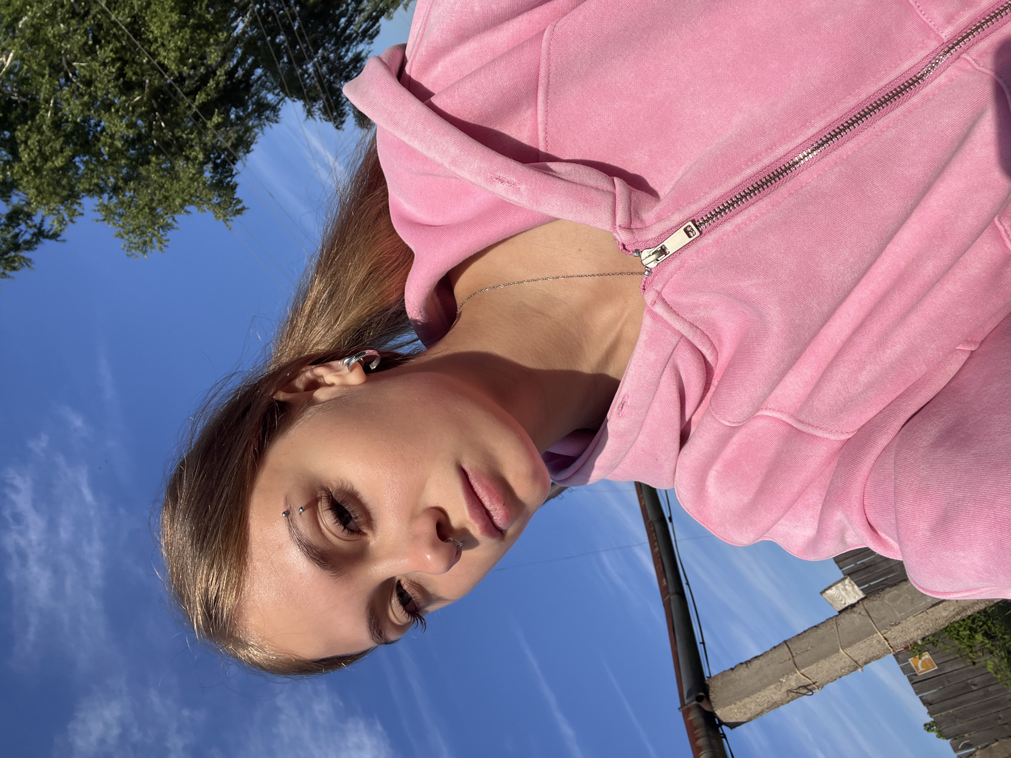 The image is a portrait of a young person, likely female, captured outdoors with a tilted perspective, making the subject appear to be looking upwards into a bright blue sky with sparse white clouds. She has light brown hair and a neutral expression, looking towards the camera.
The subject is wearing a textured pink zippered hoodie, which appears to be made of a soft material like velour or fleece. A thin silver chain necklace is visible around her neck. She has several silver piercings: a curved barbell through her right eyebrow, a small hoop in her left nostril, and an ear cuff or helix piercing on her right ear. Her eyelashes are long, and her lips have a natural appearance.
In the background, against the clear blue sky, there is green tree foliage in the upper left. On the right side, utility lines are visible, along with a dark pole, a weathered concrete beam or post, and a segment of a wooden fence. A small, indistinct yellow sign is barely visible on the fence.
The scene is illuminated by bright, direct sunlight, indicating daytime and clear, sunny weather. The activity depicted is posing for a photograph. The location context for this image is Фоки, Russia. No legible text is visible within the image.