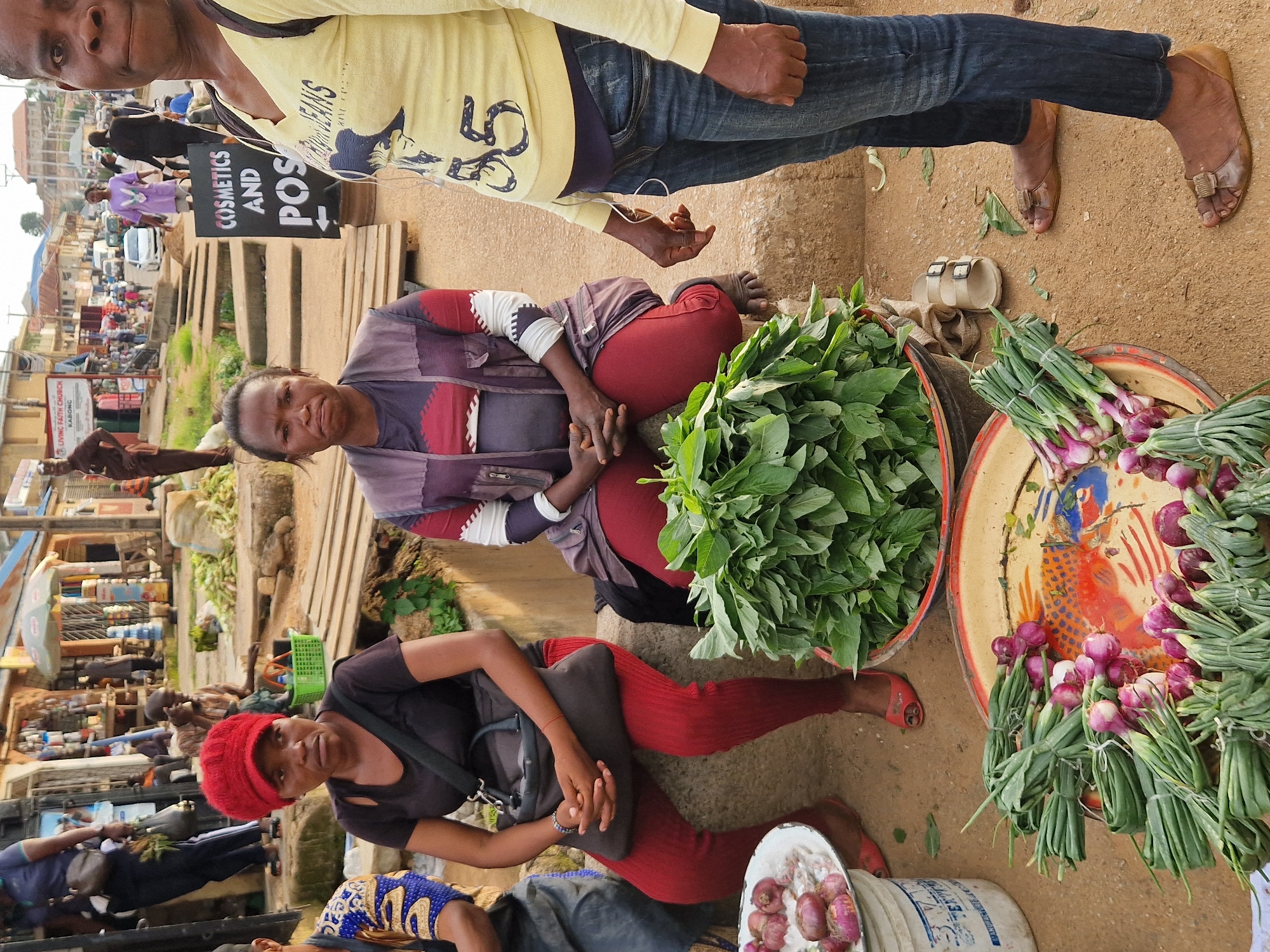 The image depicts a street market scene in Jos, Nigeria. In the foreground, three women are seated or standing near piles of fresh produce. The woman on the left wears a red headscarf and a black top, with red leggings. She is holding a dark bag on her lap. To her right, a woman in a multi-colored patterned vest and red leggings is also seated, her hands clasped. The woman on the far right, wearing a yellow top and blue jeans, stands, partially visible. Various vegetables, including leafy greens and bundles of onions or scallions, are arranged in bowls and on the ground around them. In the background, the market extends with stalls, people, and vehicles, suggesting a bustling commercial area. A sign with text including "COSMETICS AND POS" is visible. The ground is a mix of dirt and paving stones, with wooden planks and concrete structures indicating the market's layout.