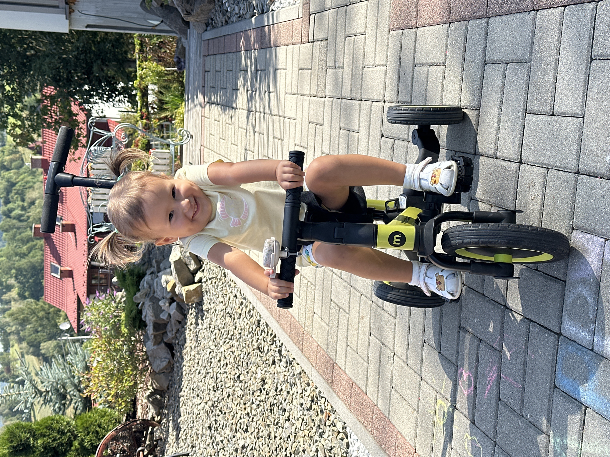 A young child with blonde hair in pigtails is riding a yellow and black tricycle on a paved walkway. The child is wearing a yellow t-shirt and shorts. The walkway is bordered by gravel on the left and a rocky area on the right. In the background, there are trees, a red-roofed house, and a hilly landscape. The scene appears to be a residential area in the city of Wisła, Poland, possibly a backyard or a front yard. The child is smiling and looking at the camera. The tricycle has three wheels and a handle. There are some chalk drawings on the pavement.