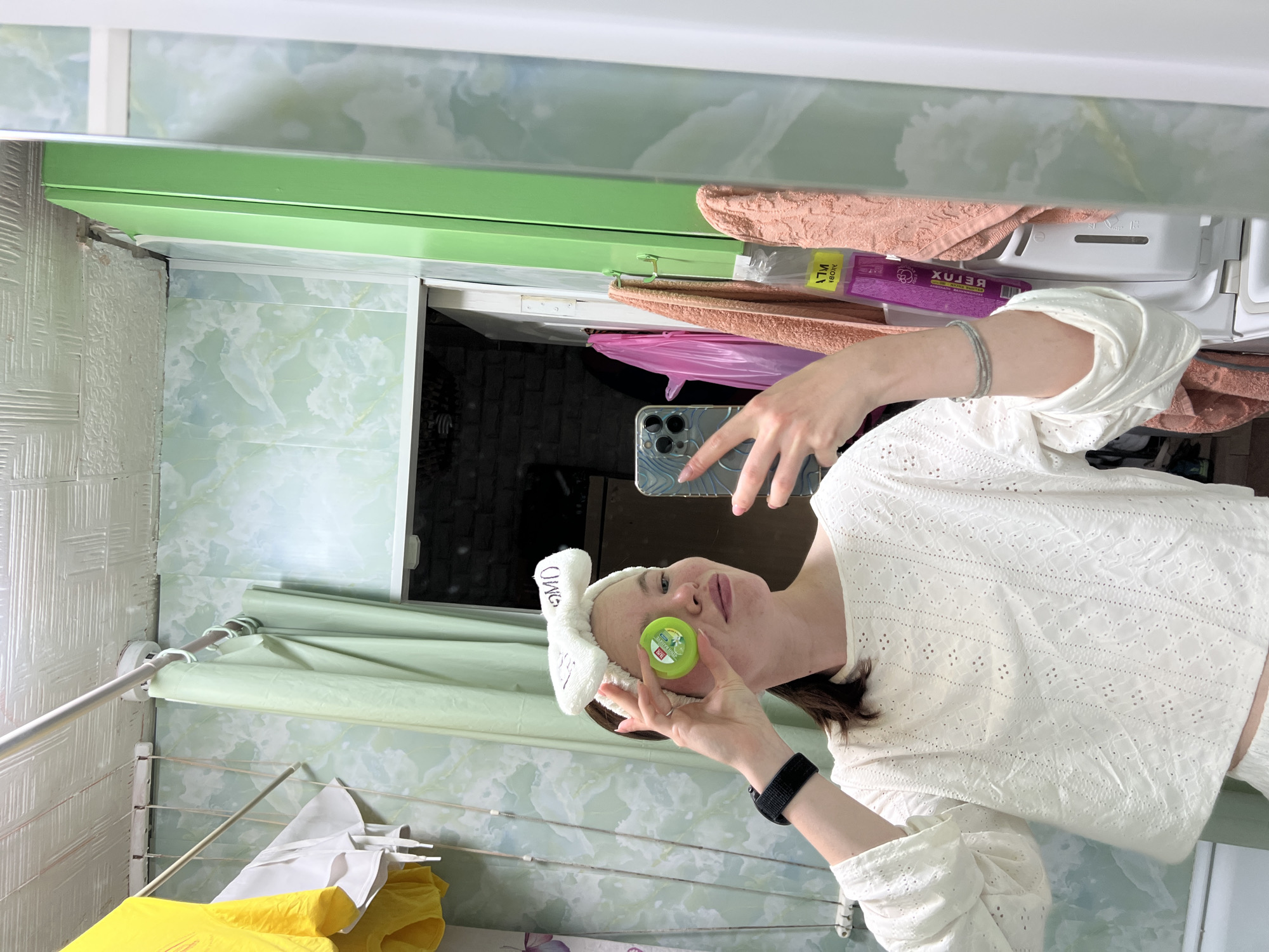 A woman is reflected in a mirror, holding a green container of skincare product near her face. She wears a white headband with "OMD" written on it and a white, lace-patterned top. She holds a smartphone in her other hand, presumably taking the photo. The background reveals a bathroom with light green, patterned tiles. A shower curtain hangs to the left. A clothes drying rack with yellow and white garments is below the shower curtain. Above the woman in the reflection, items are stored on a shelf, including peach-colored towels and a purple package labeled "RELUX". The scene is likely taken in a bathroom, given the items and background. The city of Chaikovsky, Russia, cannot be verified from the given image.