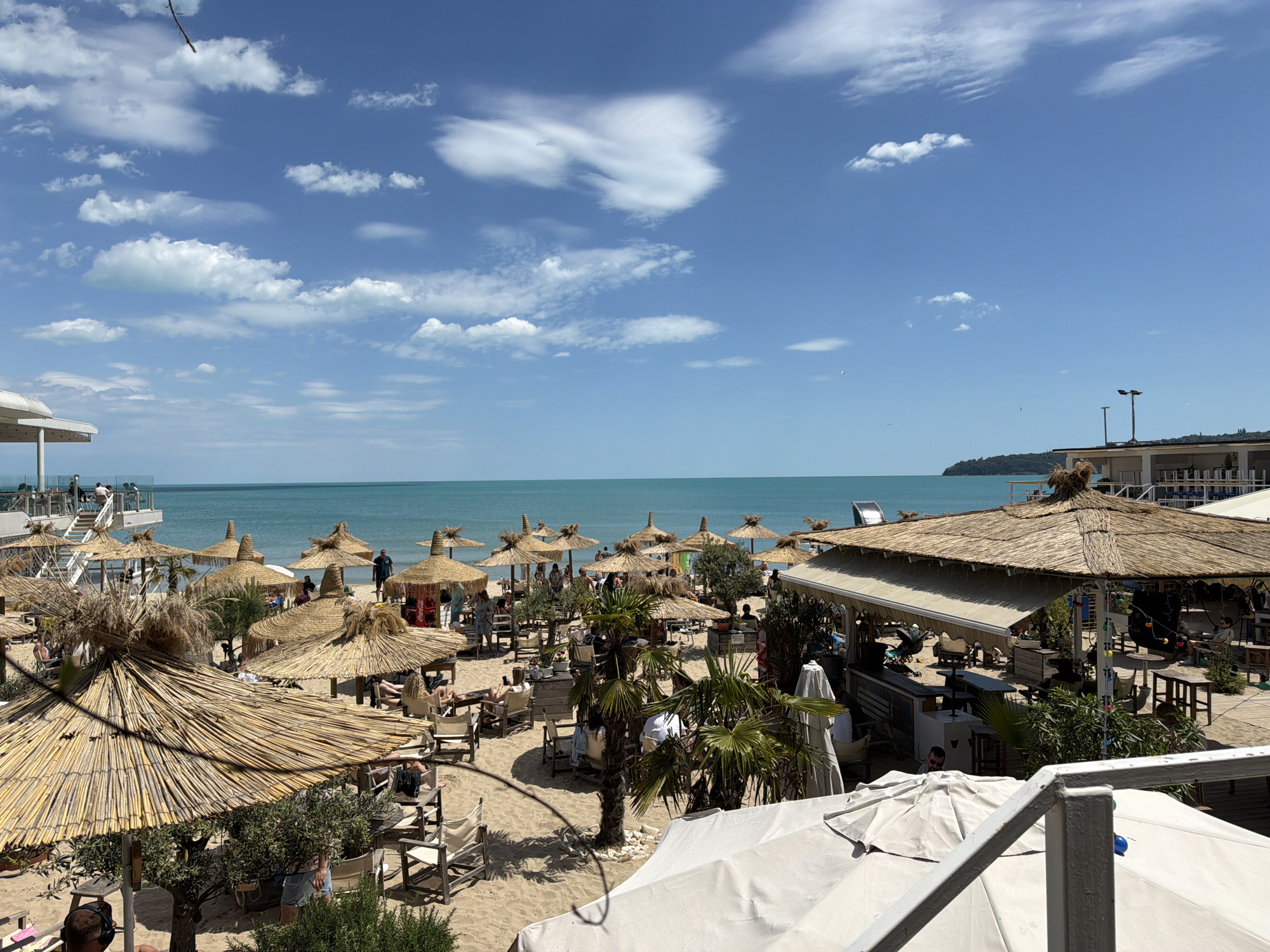 This image depicts a beach scene in Varna, Bulgaria. The foreground is dominated by several thatched-roof umbrellas providing shade to sunbathers in lounge chairs. People are visible lounging and walking about the beach. In the background is the turquoise sea, merging with a blue sky dotted with scattered clouds. Buildings and vegetation border the beach area. The scene suggests a popular tourist destination on a sunny day.