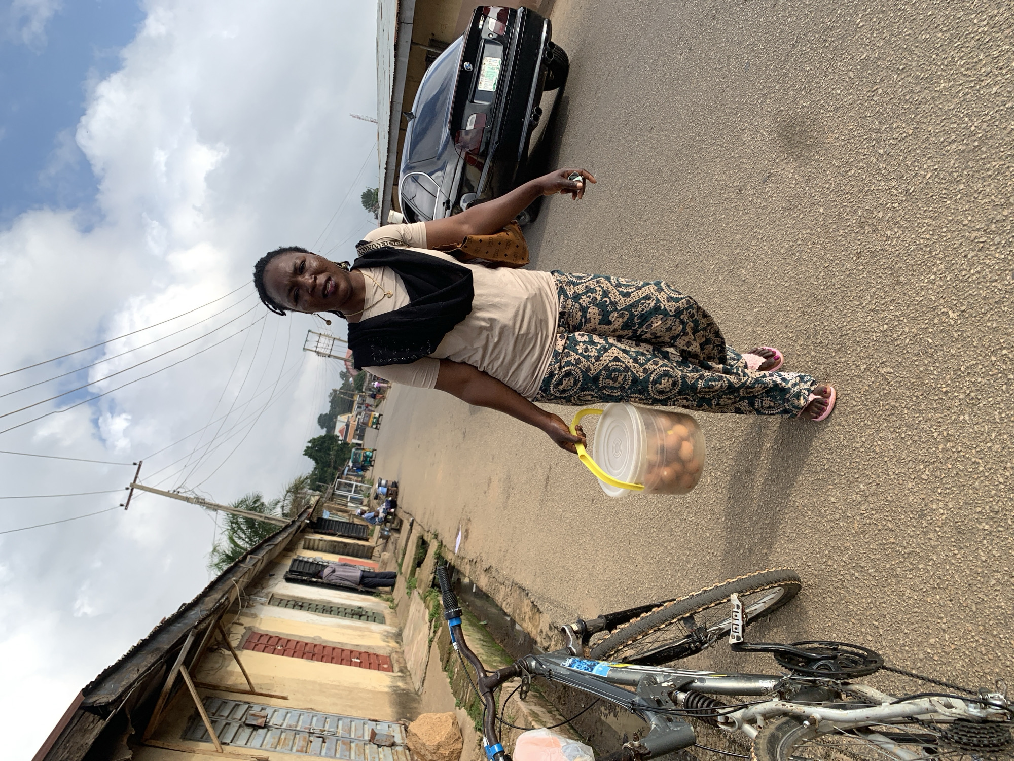 A Nigerian woman walks on a paved road in Jos, Nigeria, carrying a clear plastic bucket filled with brown eggs in her right hand. She wears a light beige t-shirt, a dark scarf draped around her neck, patterned green and gold pants, and pink flip-flops. Her left arm is bent, and a brown purse with a woven pattern is slung over her shoulder. To her left, a silver bicycle with black tires is parked on the side of the road. The woman is walking away from a row of weathered, single-story buildings with corrugated metal roofs and shuttered windows. The buildings appear to be shops, some with faded yellow and tan facades. Beyond the buildings, utility poles with numerous wires crisscross the sky. In the background, a black sedan is parked on the right side of the road, and several other vehicles, including what appear to be tricycles, are visible further down the street. The sky is cloudy with patches of blue. The road surface is rough asphalt.