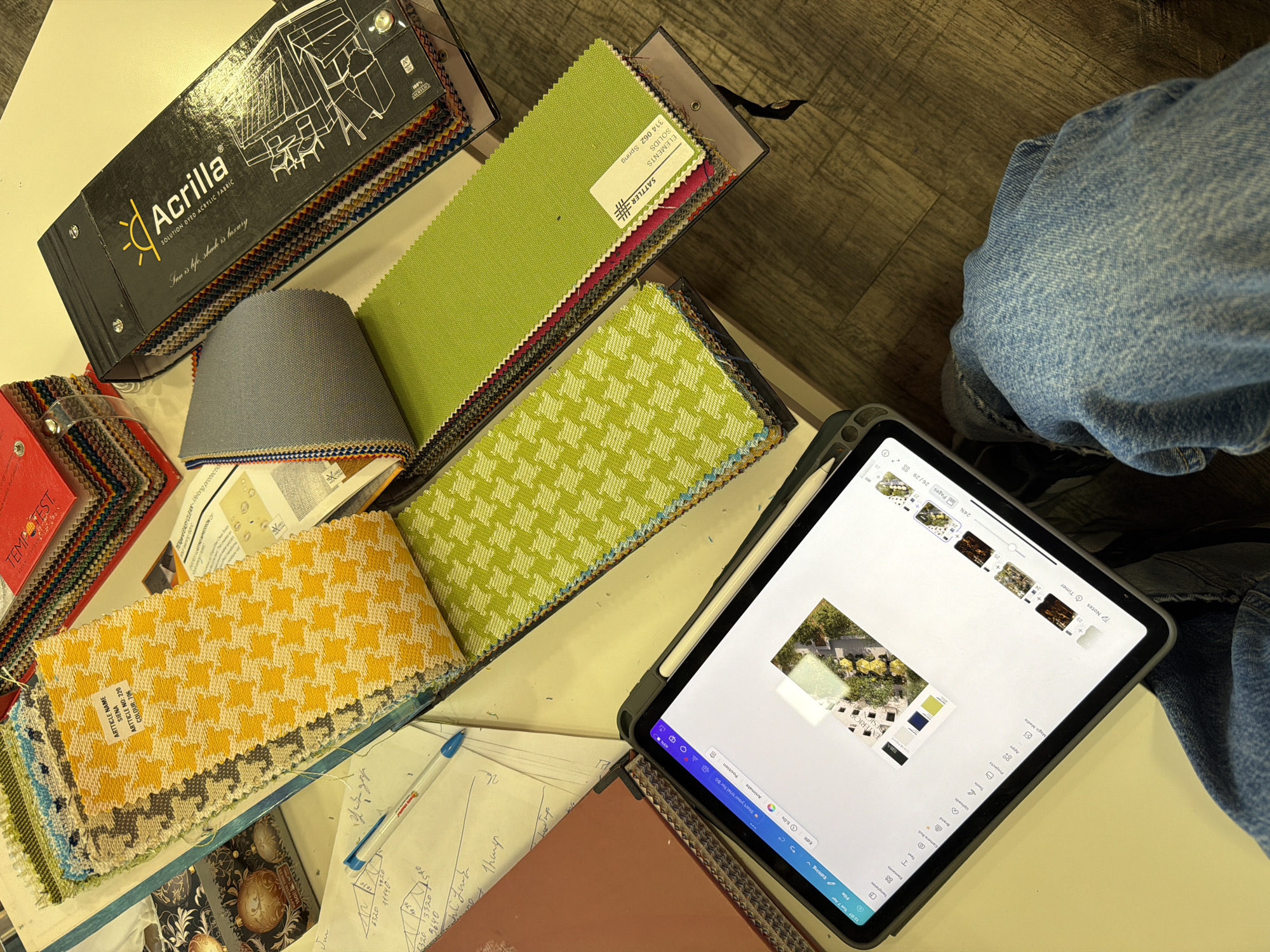 The scene presents a collection of design materials and tools spread across a light-colored surface, likely a table. A variety of fabric swatch books, featuring diverse colors and patterns, are arranged in stacks and rows. Prominent among them is a book with the "Acrilla" logo, indicating it contains samples of solution-dyed acrylic fabric. Additional swatches display houndstooth and solid green textiles. A grey fabric sample is also visible. A digital tablet rests nearby, displaying a design interface, with a project showcasing outdoor furniture arrangements, possibly for a patio or garden. Handwritten notes and calculations are scribbled on loose papers, along with a blue ballpoint pen.
Partial view of a person wearing blue jeans is visible on the right side.
The image provides no data on the location of the scene in Yerevan, Armenia.
