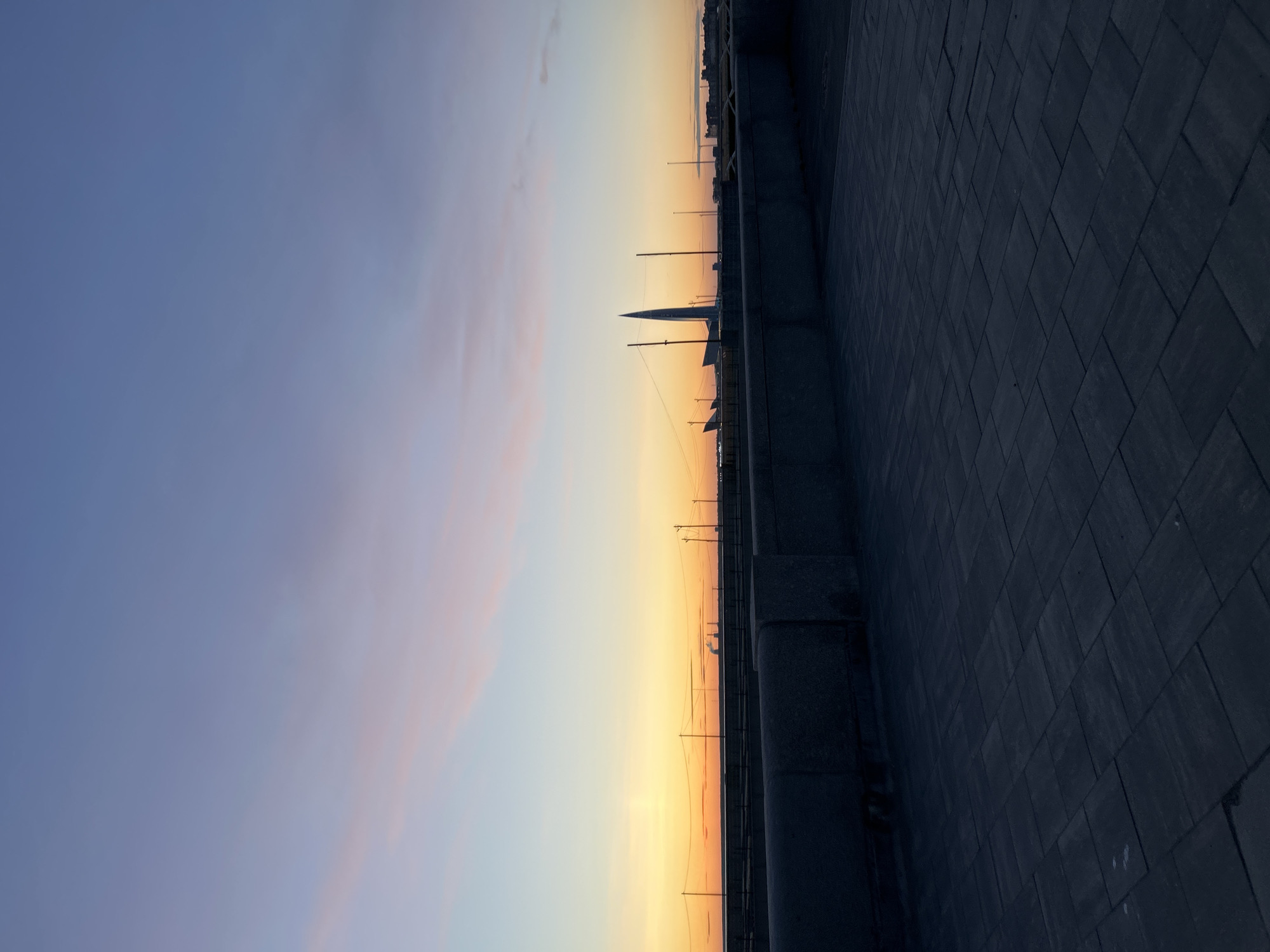 The image depicts a waterfront view in Saint Petersburg, Russia, during a sunset or sunrise. The left side of the frame is dominated by a blue and pink-tinged sky, while the right side shows a stone embankment with a tiled surface. In the middle distance, the Lakhta Center skyscraper is visible against the orange and yellow hues of the horizon, along with smaller buildings and structures along the skyline. Lampposts and various utility poles are silhouetted against the sky. The embankment surface is wet, reflecting the light.