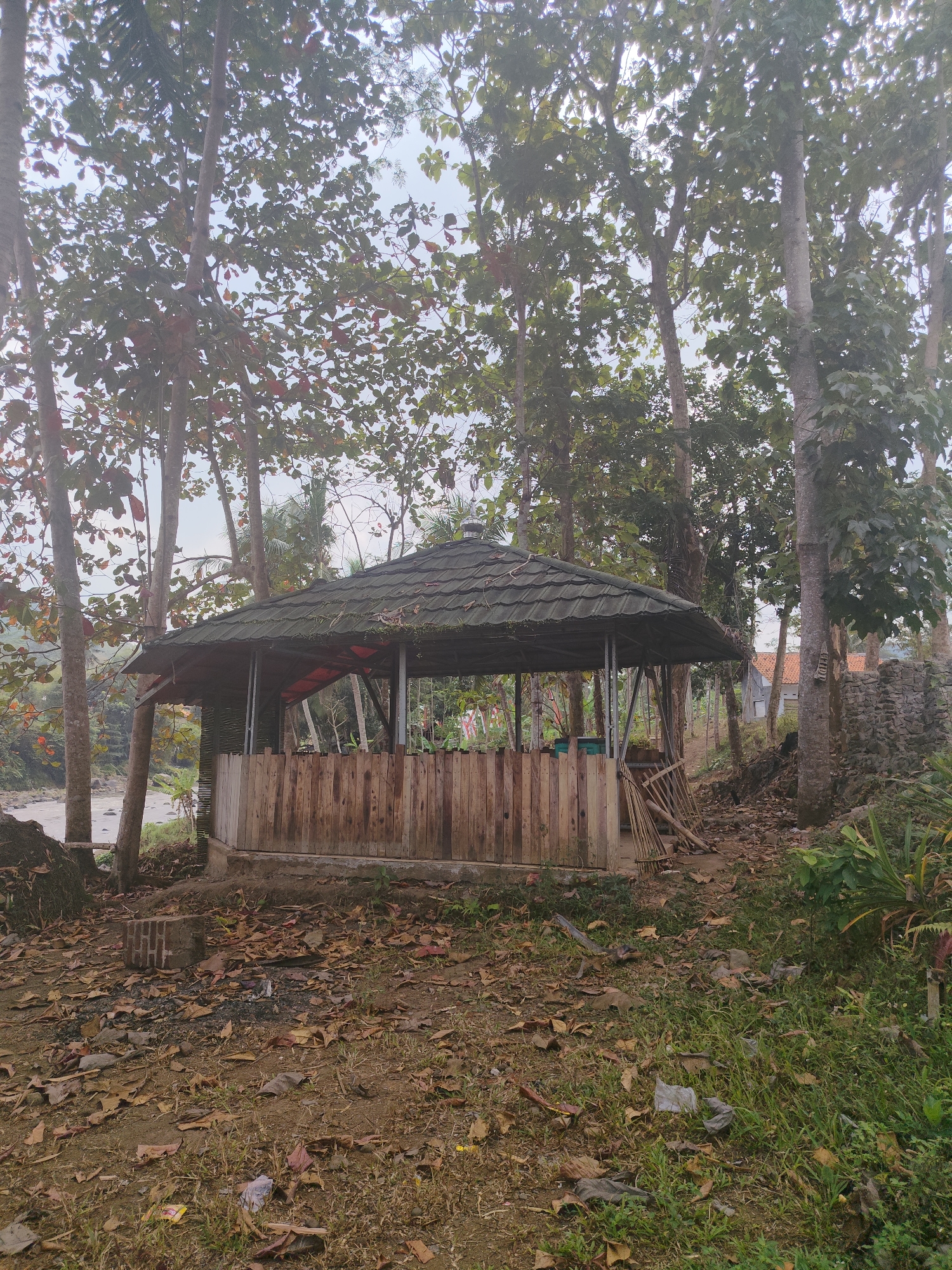The image shows an outdoor scene with a gazebo-like structure in the center, surrounded by trees. The structure has a dark, tiled roof and wooden walls. Inside, there appears to be some seating or tables, and a red tarp is visible. The ground around the structure is covered in dry leaves and some patchy grass. In the background, there are more trees, a stone wall, and a building with an orange roof. A river or body of water is visible to the left of the structure. The scene appears to be in a natural or semi-natural setting, possibly a park or a rural area. There are no people visible in the image. The lighting suggests it is daytime, possibly overcast. There is some debris and litter visible on the ground.