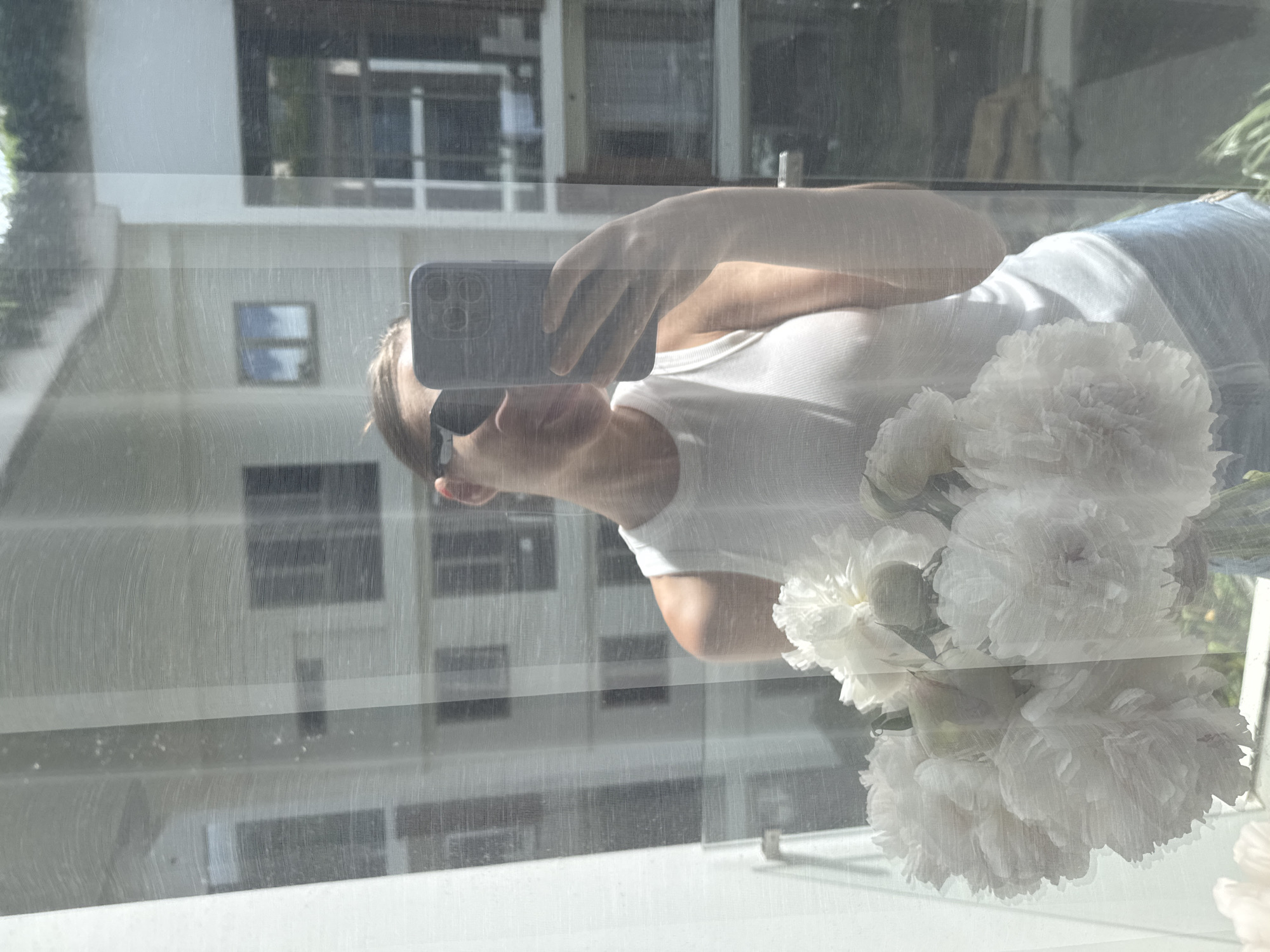 The image captures a selfie reflection of a woman in a window or glass door, viewed through a sheer, light-colored curtain or screen, in Choeng Thale, Thailand. The woman's face is partially obscured by a dark-colored smartphone, held in her right hand, which features a multi-camera lens array. She is wearing dark sunglasses, a white ribbed tank top, and light blue shorts. A bouquet of large, white, multi-petaled flowers, consistent with peonies, is positioned in the lower-left foreground. The background, visible through the sheer screen and as a reflection, consists of white multi-story residential buildings with dark-framed windows, under a bright, sunny sky. Green foliage is visible above and around the exterior buildings.