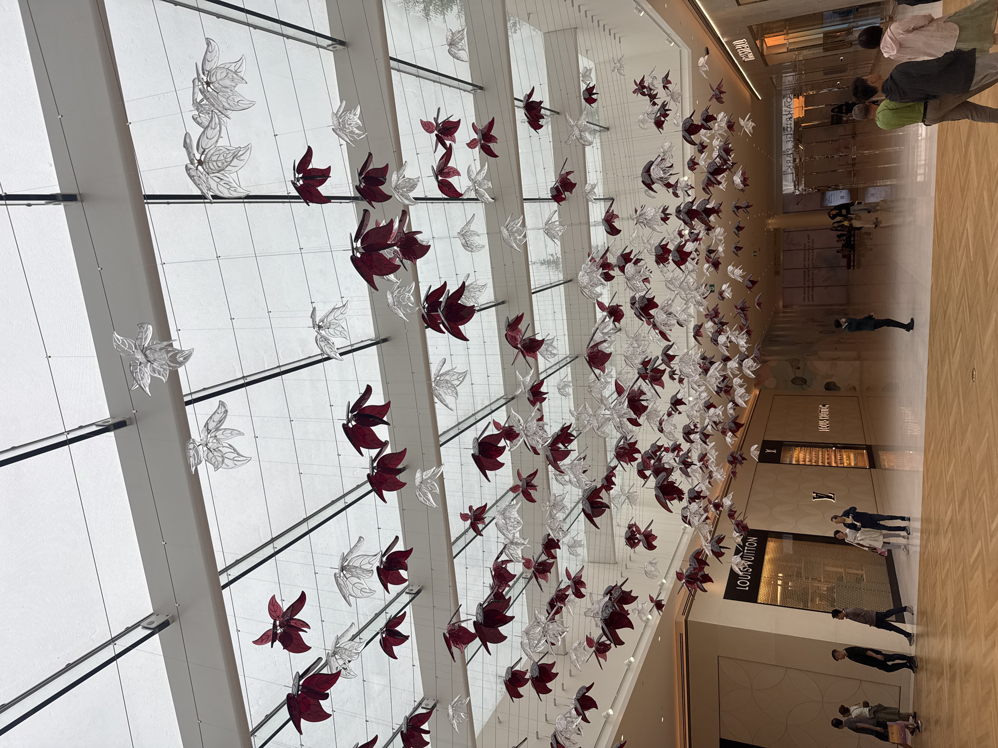 The image shows the interior of a modern shopping mall with a high ceiling constructed of glass and metal beams. Suspended from the ceiling is an elaborate art installation composed of numerous translucent and dark red leaf-shaped ornaments. The floor is polished wood. On the ground level, several people are walking and standing, some near storefronts. Prominently visible is a Louis Vuitton store with its distinctive signage. The scene suggests a high-end retail environment. Based on the Louis Vuitton store and the architectural style, this location is likely Vancouver's luxury shopping district, possibly The Fairmont Pacific Rim or a similar upscale mall.