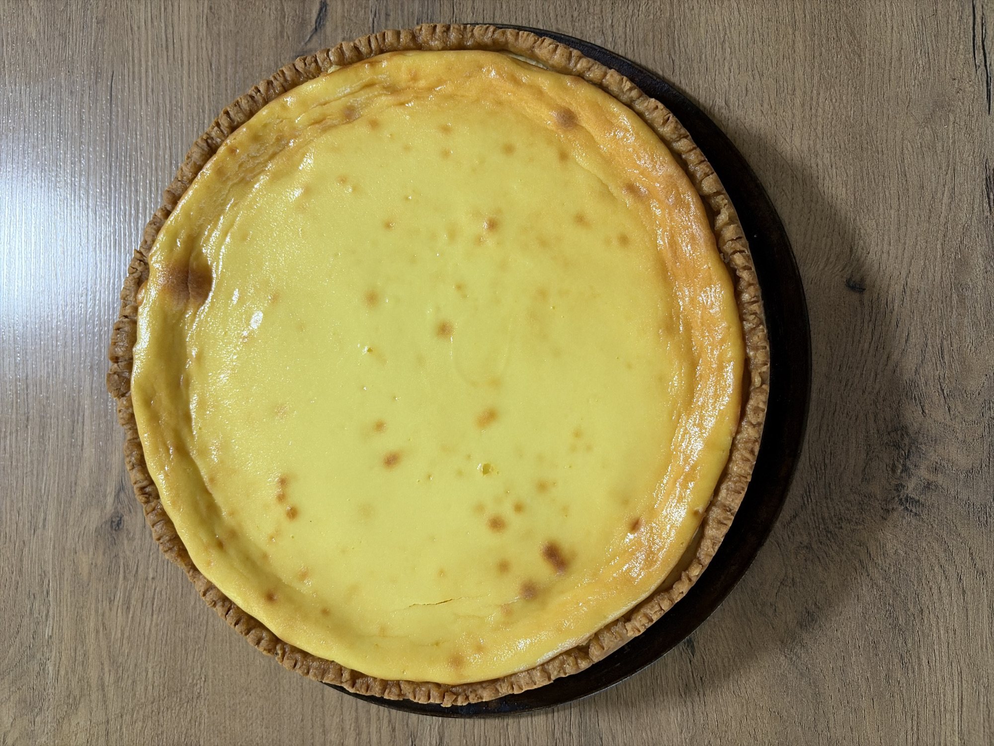 A close-up, overhead shot shows a baked cheesecake on a dark circular pie plate, resting on a light brown wooden table. The cheesecake has a golden-brown, crimped crust. The filling is a smooth, pale yellow, with some slightly darker, irregular spots on the surface, indicating it has been baked. The lighting creates some gentle highlights on the cheesecake's surface. The wooden table has a visible grain pattern. The image is cropped to focus solely on the cheesecake and the immediate surface it sits on, with no other objects or surroundings visible. There are no people or animals in the image. The scene is indoors. The location of Oxentea, Moldova is not depicted or inferable from the image.