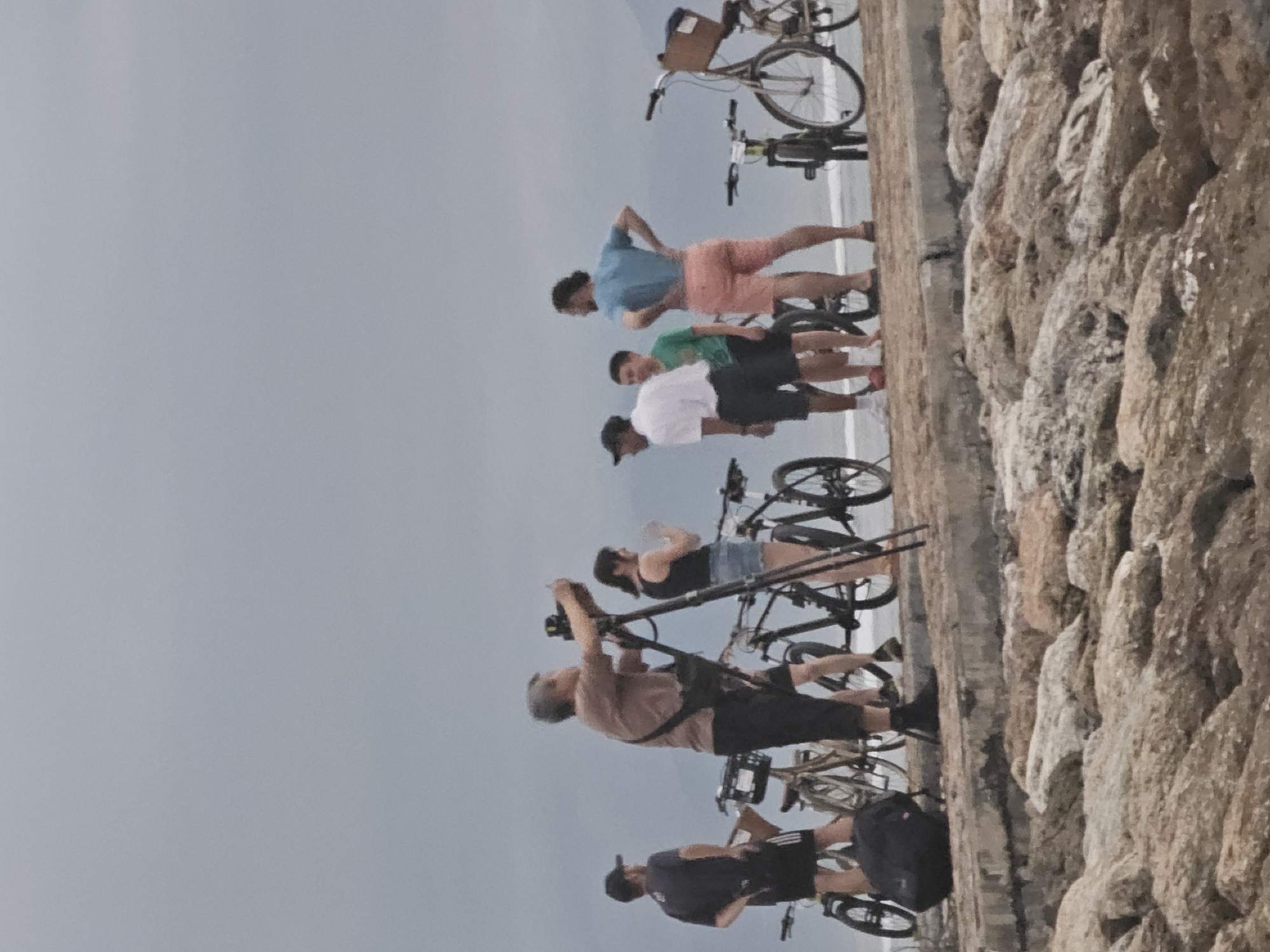 The image shows a group of people and bicycles lined up along a stone wall, presumably a breakwater or similar structure. To the right, the wall is made of large, light-colored rocks. To the left is a gray sky, suggesting an overcast day. The people are dressed in casual clothing, including shorts, T-shirts, and tank tops. A few of them are holding drinks or taking pictures with cameras. Most of the bicycles appear to be standard bicycles. The location is likely a coastal area, possibly in the vicinity of Denpasar, Indonesia.
