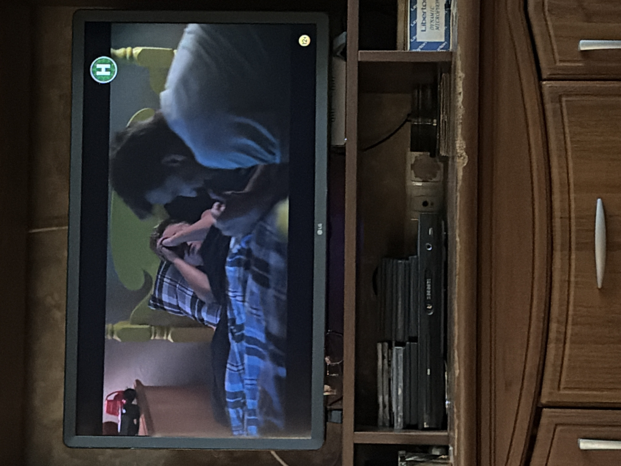 The image shows a television screen displaying a scene from a movie or show. On the screen, a person is lying in bed with their hands over their face, appearing distressed. Another person is leaning over them, possibly offering comfort or interacting with them. A red toy truck is visible on a nightstand to the left of the bed. A green circular logo with an "H" inside is in the upper right corner of the screen, and a circular icon with "12+" is in the lower right corner. Below the television, a wooden cabinet holds a DVD player, stacks of DVDs, and a box labeled "Liberton DYNAMIC MICROPHONE." The overall environment appears to be a domestic setting.