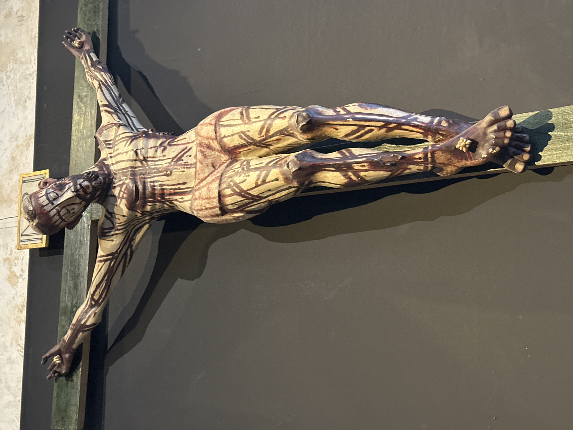 A close-up shot depicts a crucified figure, possibly a sculpture or artwork, mounted against a dark gray wall. The figure, seemingly of human anatomy, exhibits a pattern of dark lines or markings across its beige surface. The cross itself is constructed of green-hued wooden planks. Metallic embellishments are present on the figure's hands and feet, suggesting stylized crucifixion wounds. The background is a plain, dark gray wall. This picture might have been taken in a museum or religious space in San Miguel Zinacantepec, Mexico.