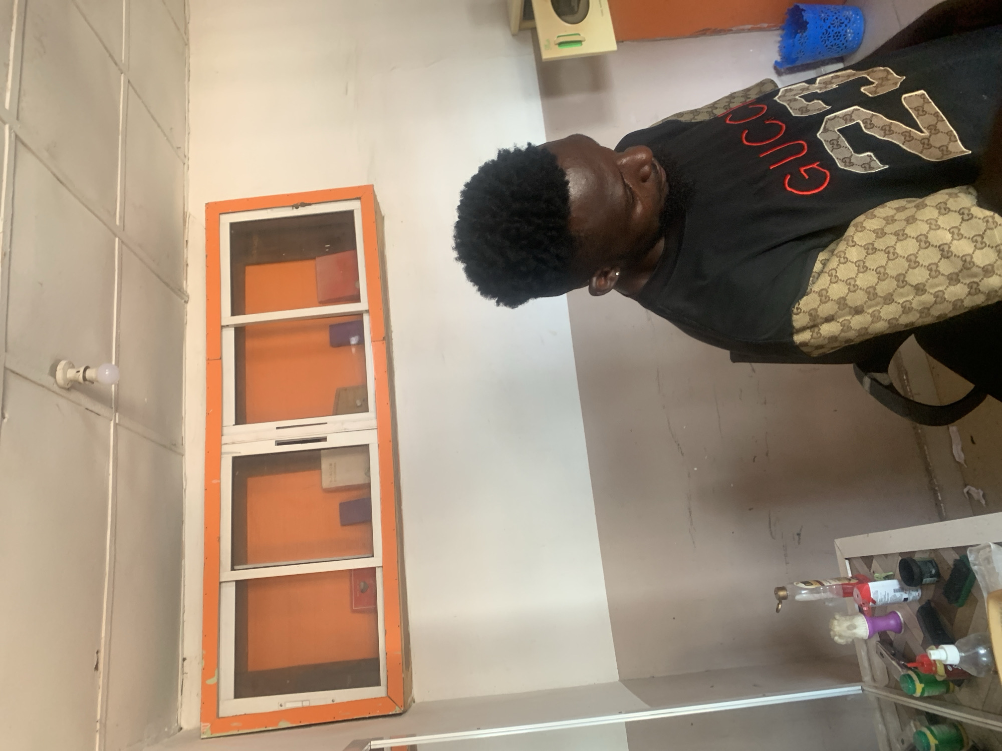 In Jalingo, Nigeria, a young man with dark skin and a black, curly hairstyle is seated in a chair. He is wearing a black t-shirt with tan sleeves featuring a repeating "GG" pattern, and the front of the shirt has "GUCCI" printed in red with "23" underneath. He is looking downwards.
The environment appears to be an indoor space, possibly a barbershop or salon. Above him, a white ceiling with visible seams and a single light fixture is present. A long, orange-framed cabinet with sliding glass doors is mounted on the wall. Inside the cabinet, several small items, some appearing to be books or boxes, are visible against an orange background.
To the left of the man, a mirror and a counter are partially visible. On the counter, several bottles and containers, likely hair products or grooming supplies, are arranged. To the right of the man, a portion of a light-colored appliance or cabinet is visible, along with a blue plastic basket. The walls are painted in two tones, with a lighter color above a darker, grayish-brown section towards the bottom. The floor is partially visible and appears to be tiled.