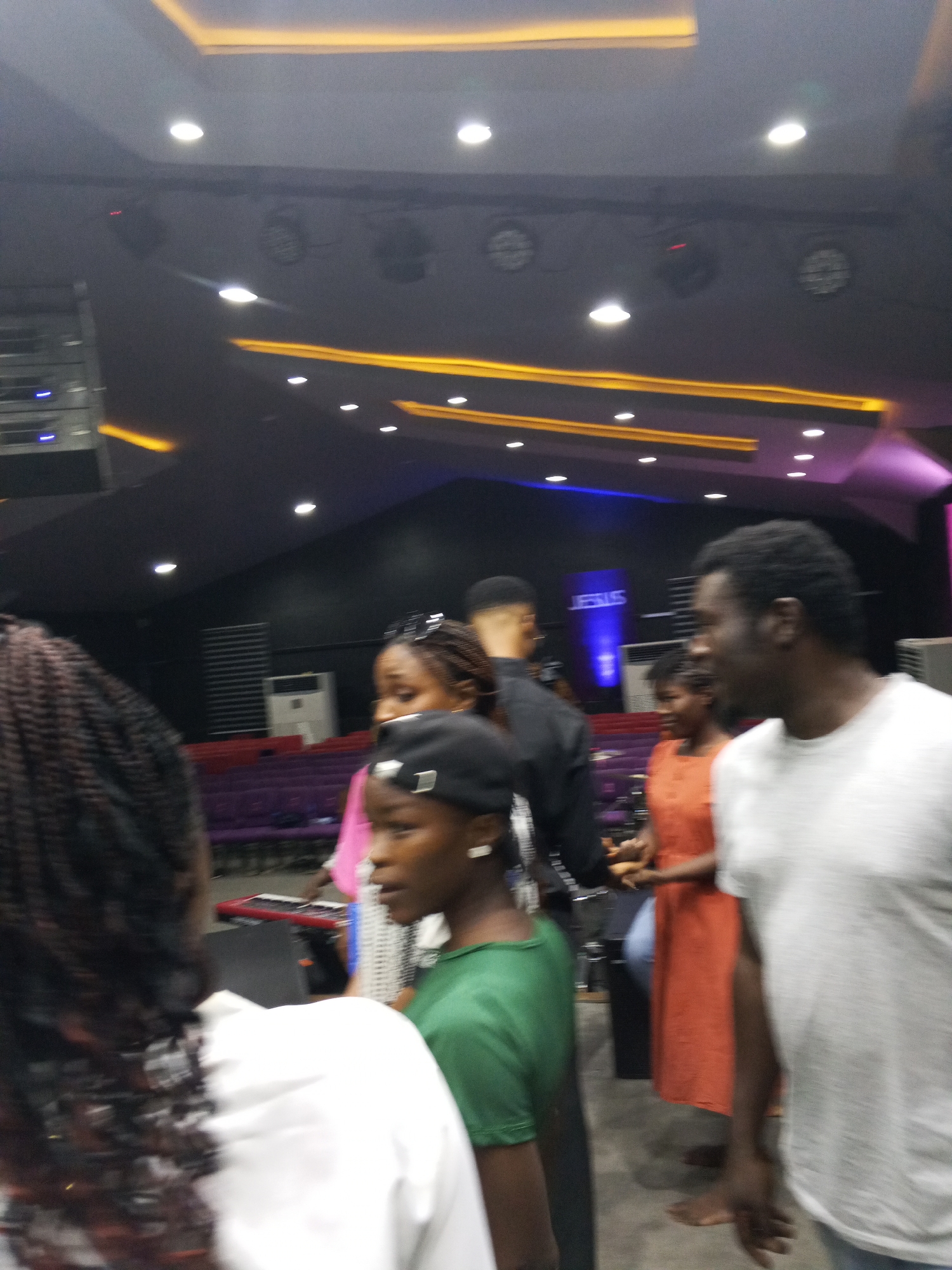 The image captures a blurred scene inside what appears to be a church or auditorium, evidenced by the presence of stage lighting, a sign displaying "JESUS," and rows of purple seats in the background. A group of people is present, with one man wearing a white t-shirt in the foreground. Other people are wearing a green shirt and a black shirt, and someone wearing a coral dress is visible further back. The image quality is poor, and the location is not possible to determine only from the image alone, but I believe it might be in Abuja, Nigeria.