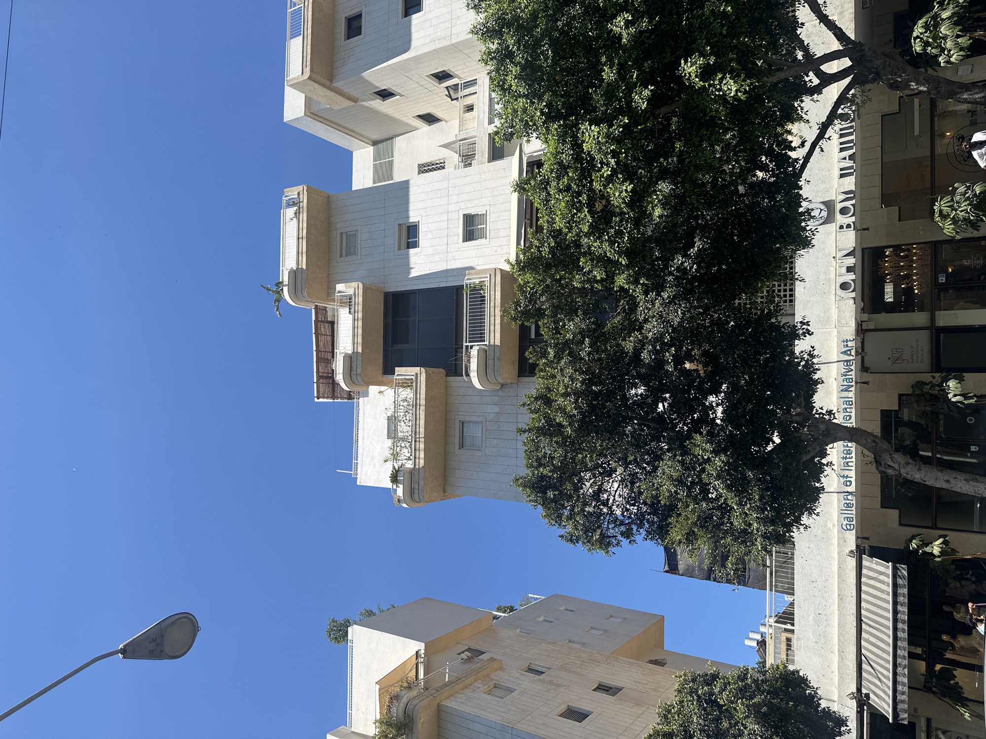 The image shows a street view in Tel Aviv, Israel, with modern white buildings dominating the scene. A clear blue sky is visible above. In the foreground, lush green trees obscure part of the lower floors of the buildings. One building features a sign that reads "Gallery of International Naive Art" and "JOHN BOΥ ΠΑΠΠΑΣ" in blue lettering. Shops with awnings are visible on the ground floor. A lamppost extends from the upper left corner of the frame. The architecture suggests a contemporary urban environment.