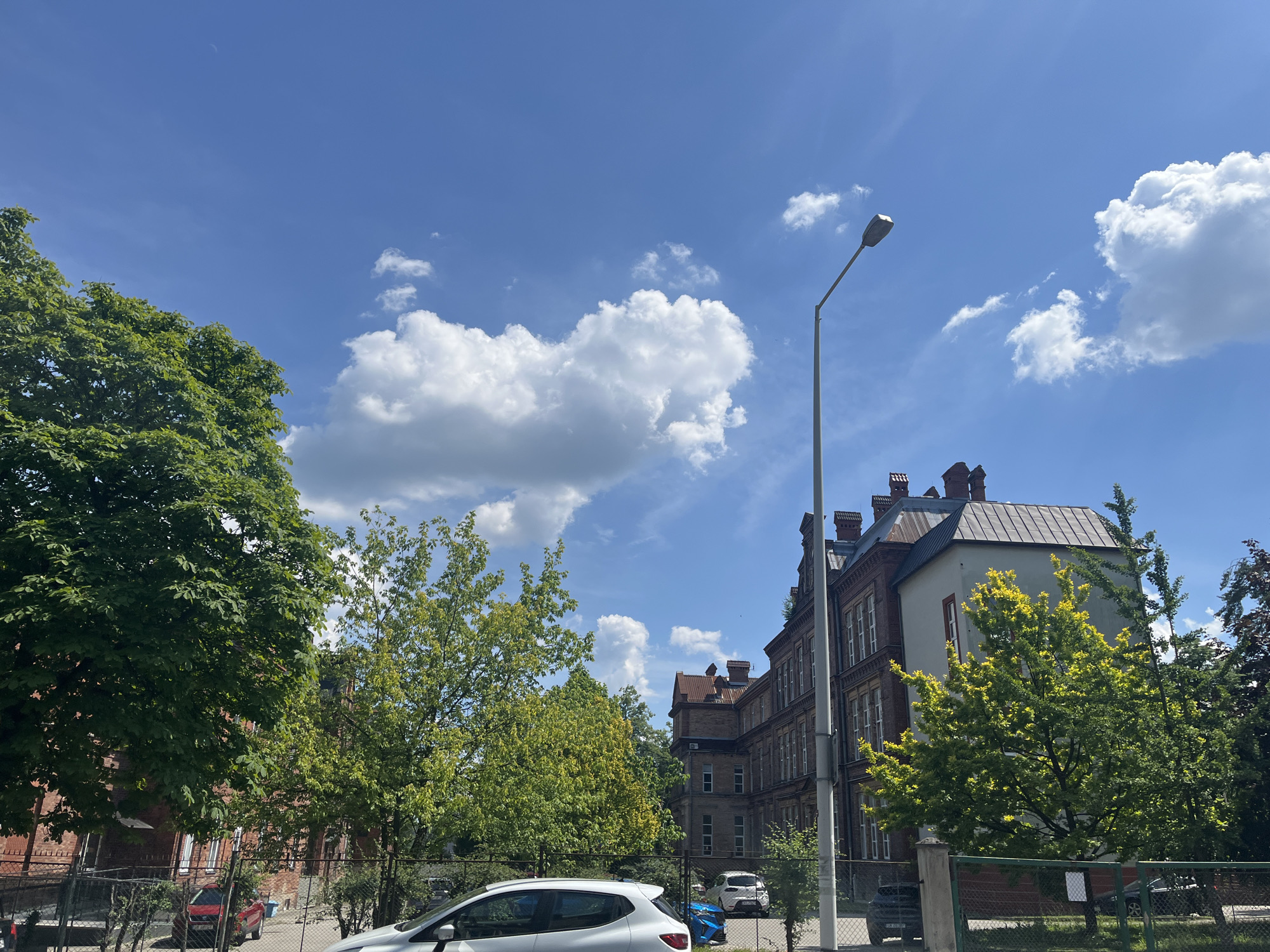 The image depicts an outdoor scene with a bright, blue sky dotted with fluffy white clouds. A tall, modern street lamp stands on the right side of the frame. In the middle ground, an old, multi-story brick building with numerous windows and a dark roof is visible. To the left of this building, lush green trees with dense foliage are prominent. A chain-link fence runs along the foreground, separating the viewer from the scene beyond.
Several cars are parked or driving on a paved surface behind the fence, including a white hatchback in the immediate foreground, a dark blue car, and a red car further back. The sun appears to be high in the sky, suggesting it is daytime. The weather seems fair and clear, with the sun casting shadows and illuminating the scene. The overall impression is a view of an urban or suburban environment with a mix of older architecture and greenery. There is no visible text in the image.