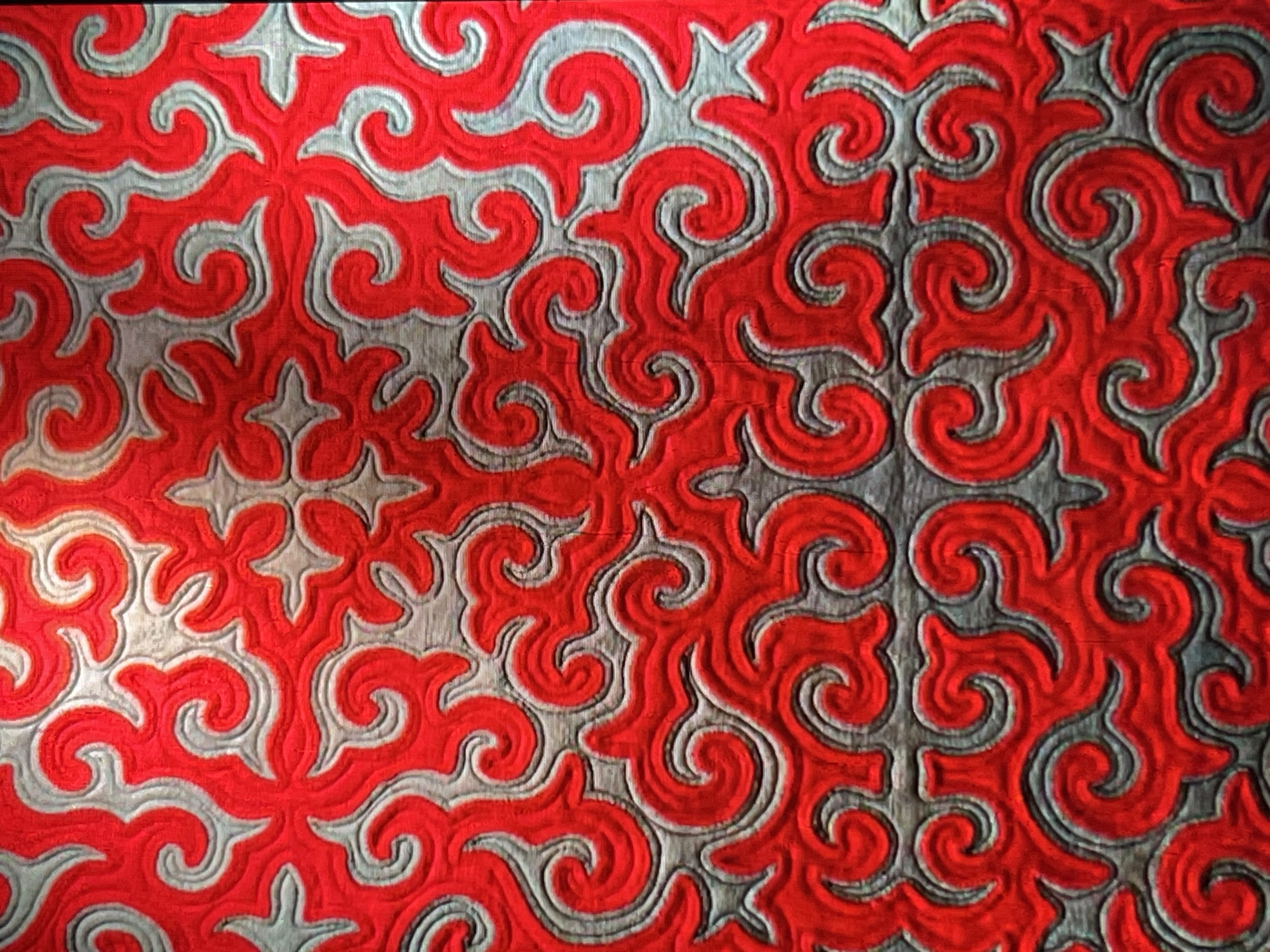 This is a close-up, eye-level, full-frame shot of a red and grey patterned fabric. The pattern consists of swirling, organic shapes that are outlined in grey and filled with a textured, bright red material. The fabric appears to be a quilt or a similar textile with raised stitching or appliqué work. The lighting is even, highlighting the texture and depth of the pattern. There are no people or recognizable landmarks visible, so it is not possible to determine its location within Bishkek City, Kyrgyzstan. The image is purely a detailed examination of a textile pattern.