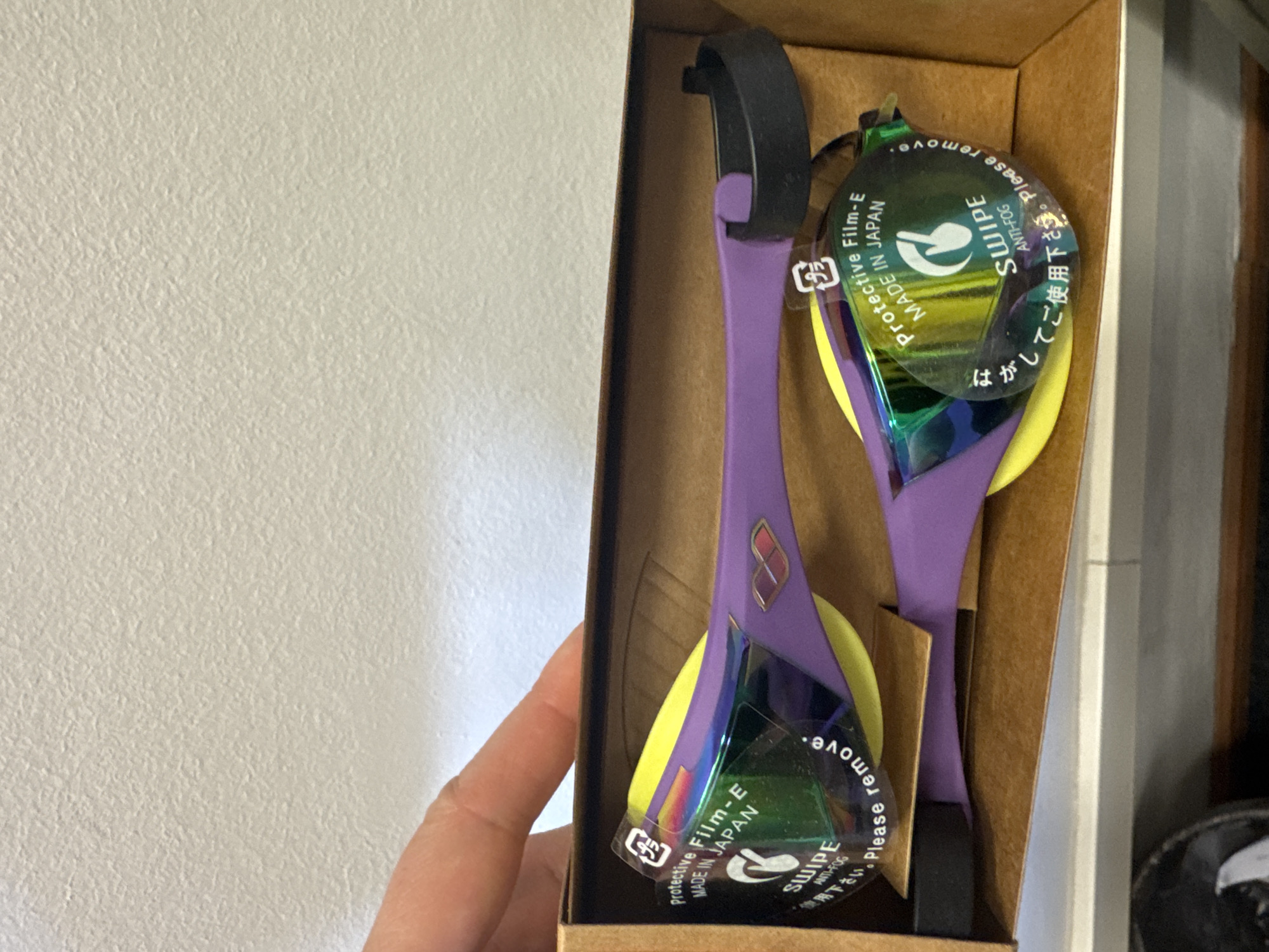 This media file captures a pair of new swimming goggles presented in a brown cardboard box, seemingly during an unboxing or preparation for use. The goggles feature a striking design with purple frames and bright yellow inner eye cushions. Their lenses are iridescent and mirrored, reflecting a range of greens, blues, and purples. A black adjustable strap is visible at the top.
Each goggle lens is covered by a clear, circular protective film. This film displays several lines of text, including "Protective Film-E," "MADE IN JAPAN," "SWIPE," and "ANTI-FOG." It also states "Please remove." and features Japanese text, "はがしてご使用下さい" (Hagashite go shiyou kudasai), which means "Please peel off and use." A recycling symbol indicating PP (Polypropylene) is also visible on the film.
A human hand with light skin tone is partially visible on the left side of the frame, holding the cardboard box. The background is a plain, light-colored (off-white) wall with a subtle textured finish, suggesting an indoor setting. The image was captured in Sofia, Bulgaria, highlighting new, Japanese-made swimming gear.