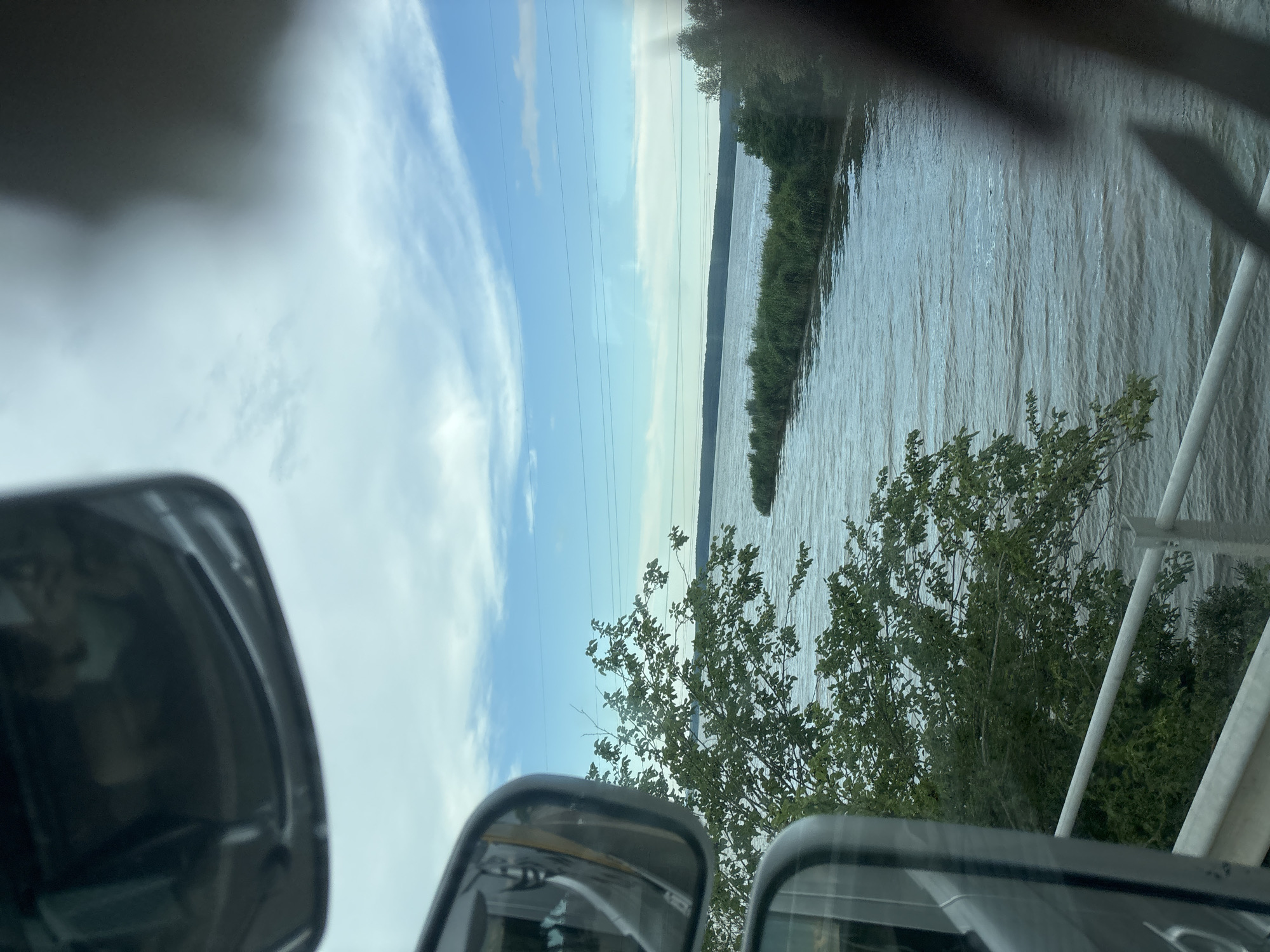 The image shows a view from a vehicle, likely a bus or truck, looking out towards a body of water. Two side-view mirrors are visible, one at the top and another on the left side, reflecting parts of the vehicle's interior and exterior. The landscape beyond the mirrors features a wide expanse of water, appearing to be a river or a lake, with vegetation lining its banks. In the background, a distant treeline is visible under a sky with scattered clouds. Power lines are strung across the sky. The image's perspective suggests movement, as if captured while the vehicle is traveling. There are no people clearly visible in the scene. The overall impression is one of a scenic journey through a natural landscape, potentially near a waterway. The specific location in the city of Konstantinovo, Bulgaria, is not discernable from the visual information provided in the image.