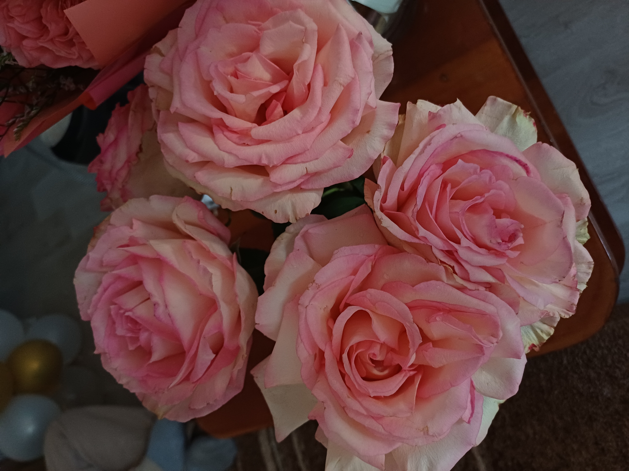 A close-up, overhead shot shows a bouquet of five light pink roses on a brown surface. The roses are in full bloom and have delicate, layered petals with darker pink edges. Some petals appear to have water droplets on them. In the background, out of focus, are balloons in shades of white and gold, and a glimpse of a pink bouquet wrapper. The scene is dimly lit, casting soft shadows.