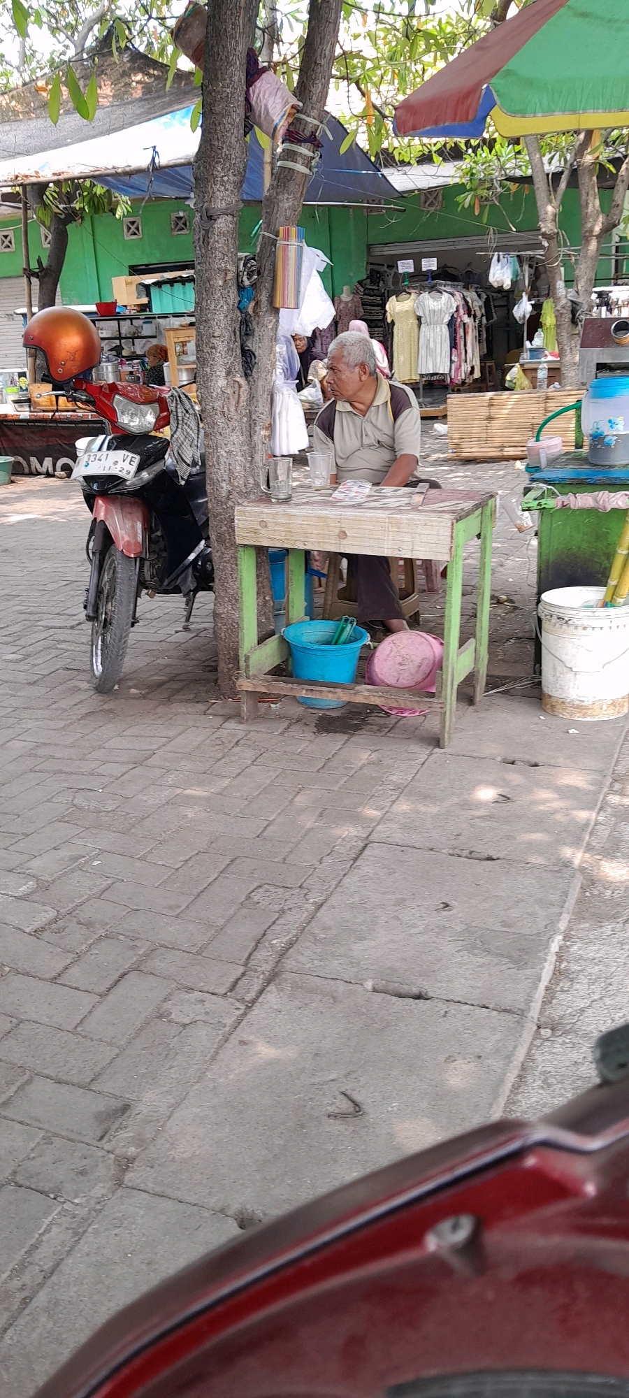 The image depicts an outdoor scene, likely a street market or roadside stall, in Pasuruan, Indonesia. In the foreground, the lower portion of a red vehicle is visible. To the left, a red and black motorcycle is parked, with an orange helmet resting on its handlebars. A man with gray hair, wearing a light brown shirt with a darker collar and dark pants, is seated at a small, light green wooden table. The table has a checkered pattern on its surface and two glasses are placed on it. A blue bucket and a pink lid are visible beneath the table. In the background, a green building with a corrugated metal roof is visible. Clothes are displayed on racks in front of the building, suggesting it may be a clothing stall. Several umbrellas, in green and red colors, are also present, providing shade. The ground is paved with interlocking concrete blocks. The overall impression is that of a bustling, everyday scene in an Indonesian city.