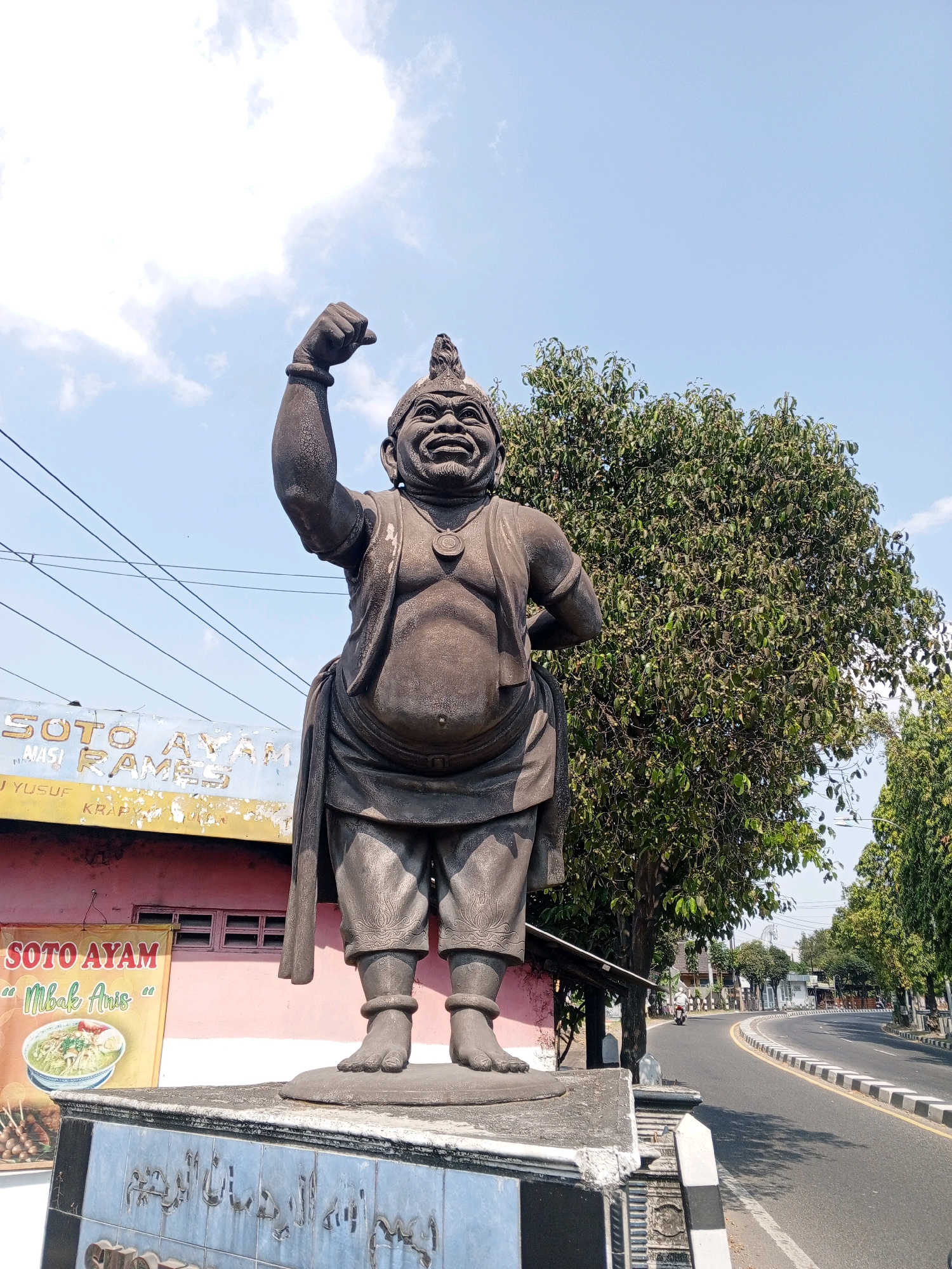 In Sewon, Indonesia, a bronze statue of a stout, smiling figure stands in front of a building that appears to be a food establishment. The statue depicts a character with a prominent belly, wearing a vest, baggy pants, and a headpiece, with one arm raised in a clenched fist. Behind the statue, a road curves to the right, flanked by trees and a sidewalk with a white and black border. A few power lines are visible in the sky. The building behind the statue has signs that read "SOTO AYAM" and "MBAK ANIS," indicating it is likely a restaurant serving chicken soup. The overall scene suggests a public space, possibly a roadside attraction or a local landmark.